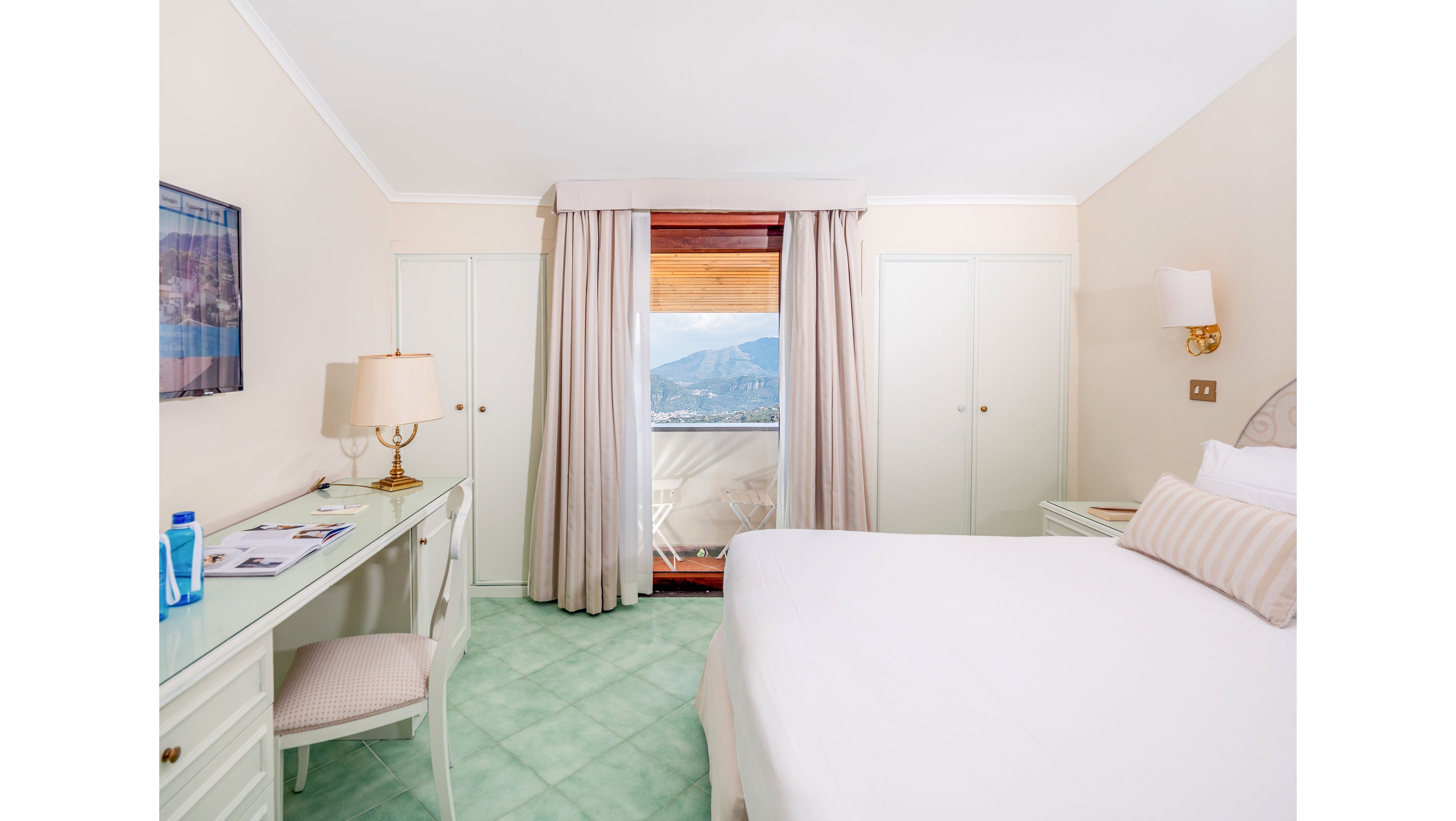 A bright hotel bedroom features a large neatly made bed with striped pillows and a soft headboard. Opposite the bed, a white desk with a lamp, chair, and scattered papers sits against the wall. Floor-to-ceiling curtains frame a glass door that opens to a balcony overlooking distant mountains and a lake. Soft pastel colors, mint green flooring, and warm lighting create a calm, inviting atmosphere.