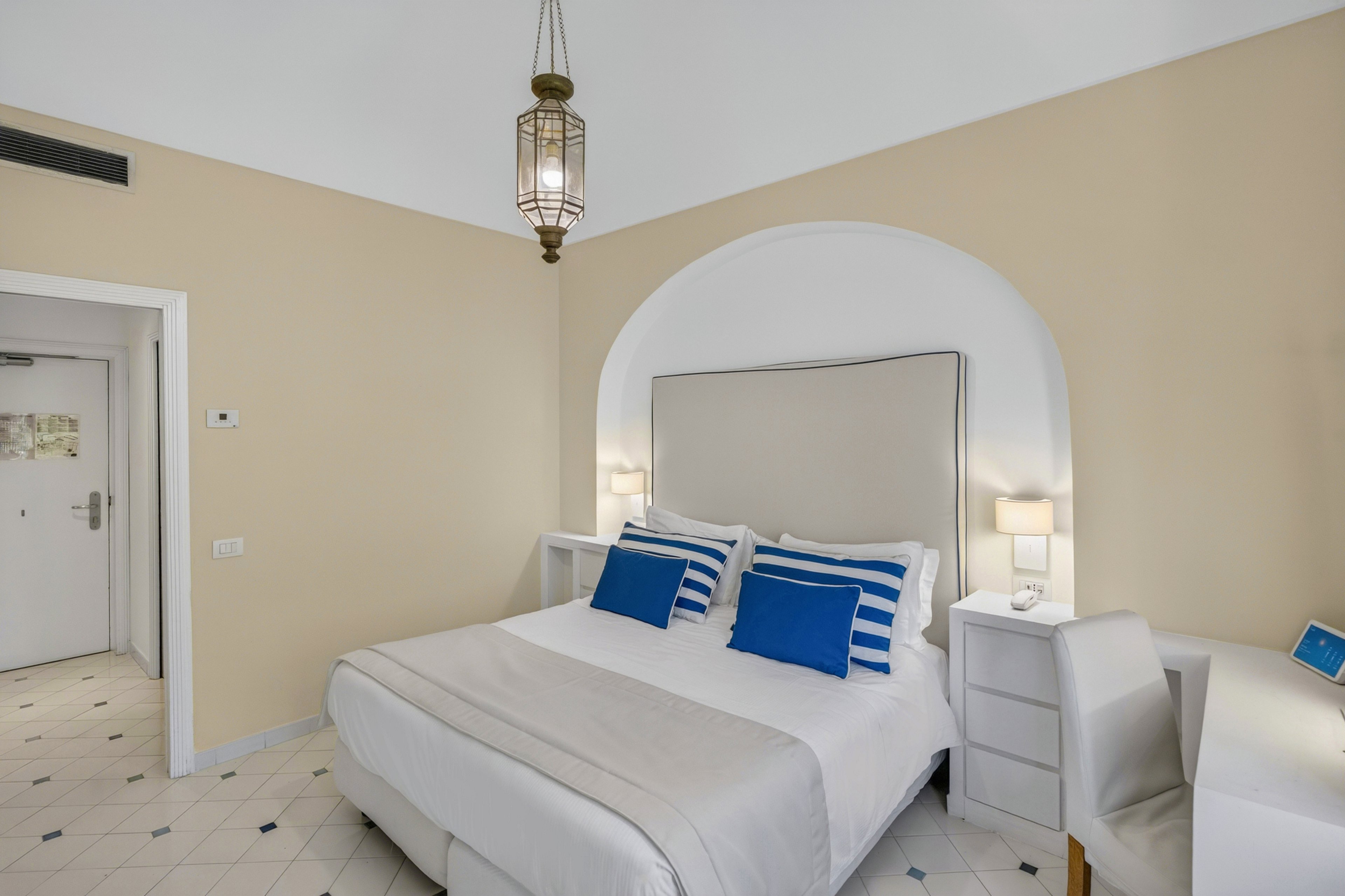 A neatly made double bed with a tall upholstered headboard is the focal point of this bright hotel bedroom. The room features white furniture, including bedside tables with lamps and a small desk with a chair. Blue accent pillows and a blue notebook add a coastal touch against the neutral walls and light tiled floor. A decorative hanging light fixture and an arched niche above the bed complete the clean, contemporary look.