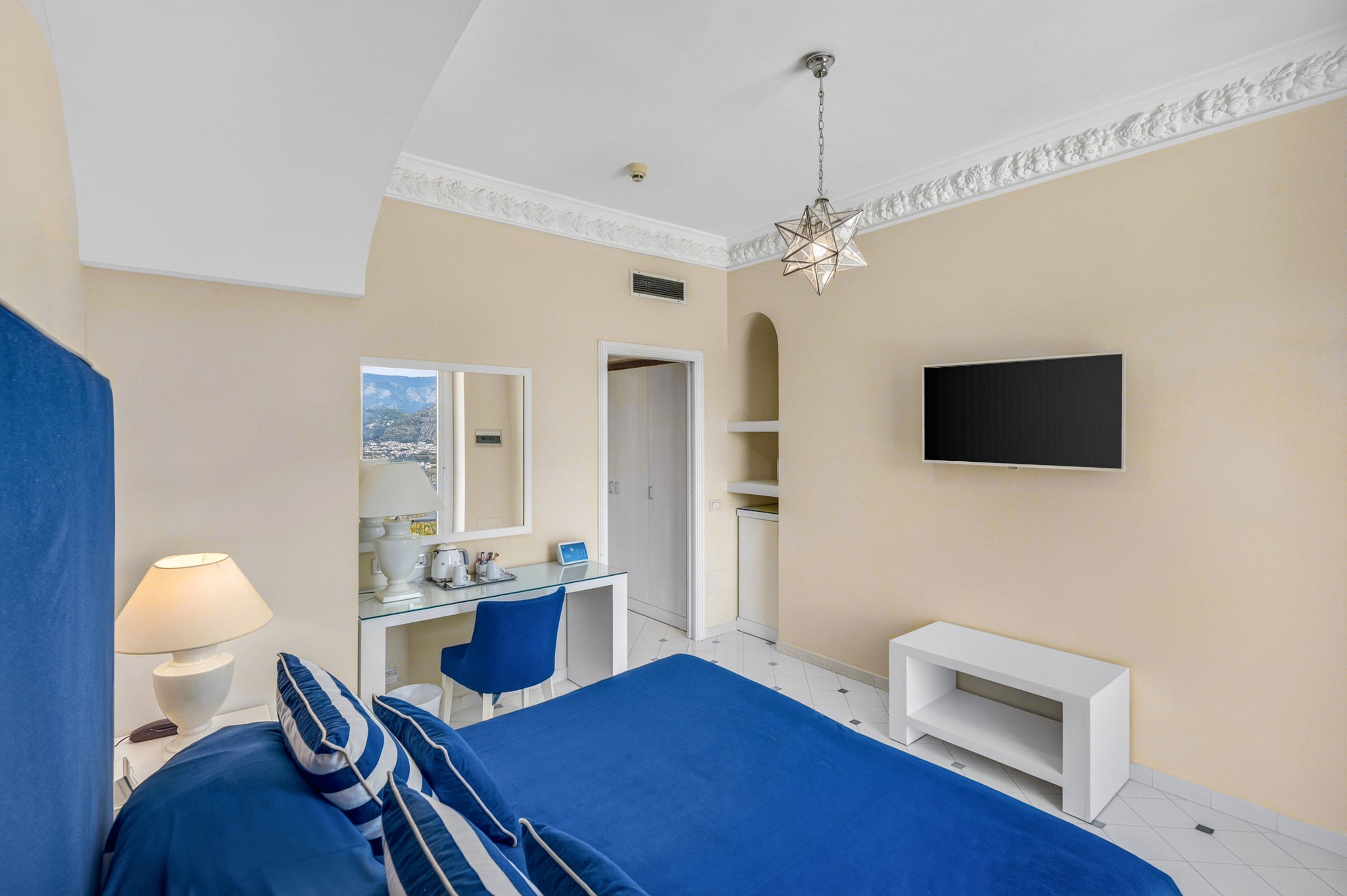 A neatly made bed with bold blue linens and striped pillows dominates this light-filled hotel room. The space features a wall-mounted flat-screen television, a white console, and built-in shelves against soft beige walls. A small vanity desk with a blue chair, mirror, and decorative items sits near the doorway, while a lit table lamp adds warmth. Ornate ceiling molding and a star-shaped pendant light contribute to the elegant, coastal-inspired style.