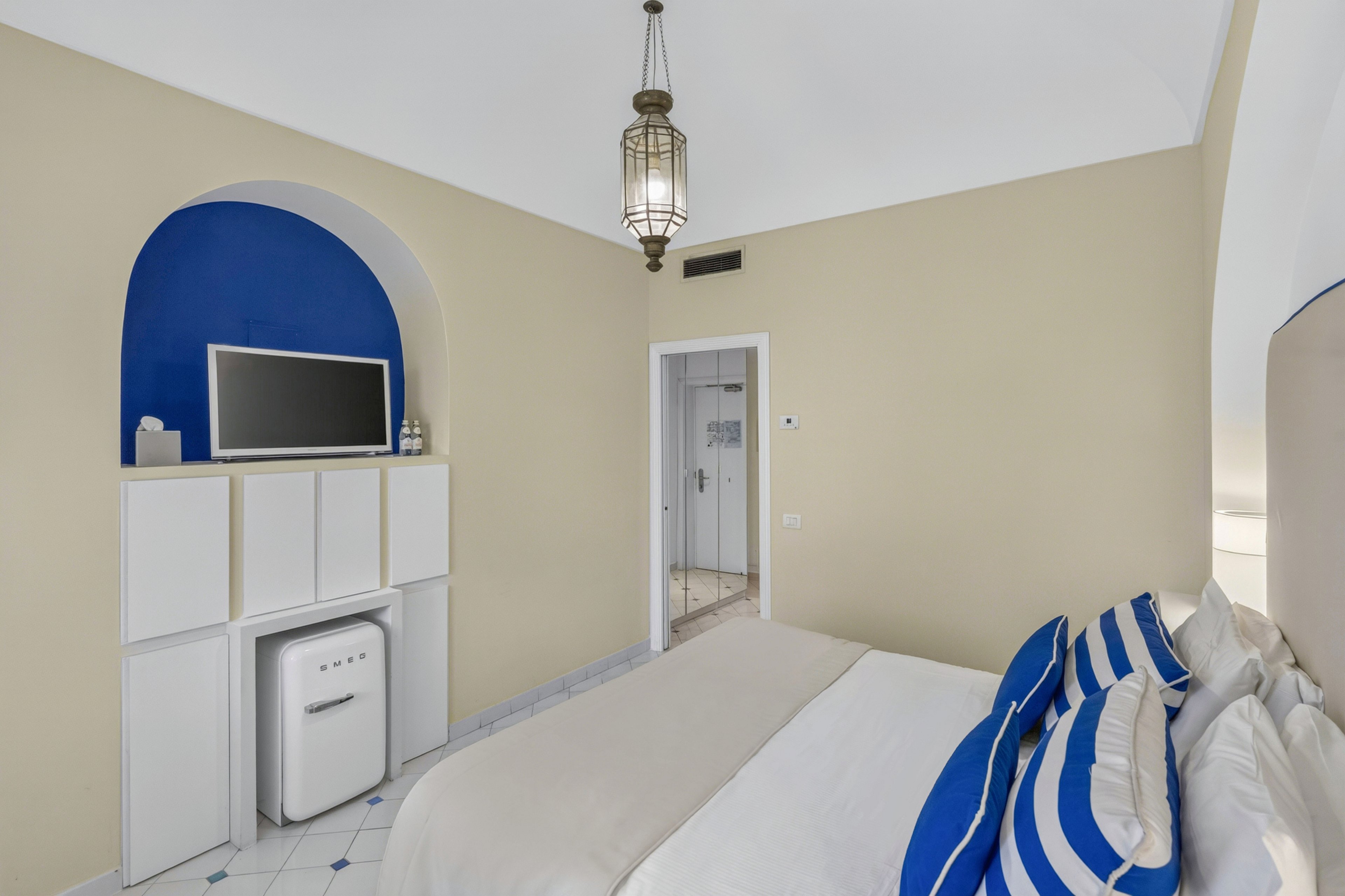 A neatly made bed with blue and white striped pillows sits in a bright hotel bedroom. A flat-screen television is mounted within a blue arched niche above white storage cabinets and a compact refrigerator. The room features light-colored walls, tiled flooring, and a hanging lantern-style ceiling light. A white door leads to an adjoining space, enhancing the clean and airy atmosphere.