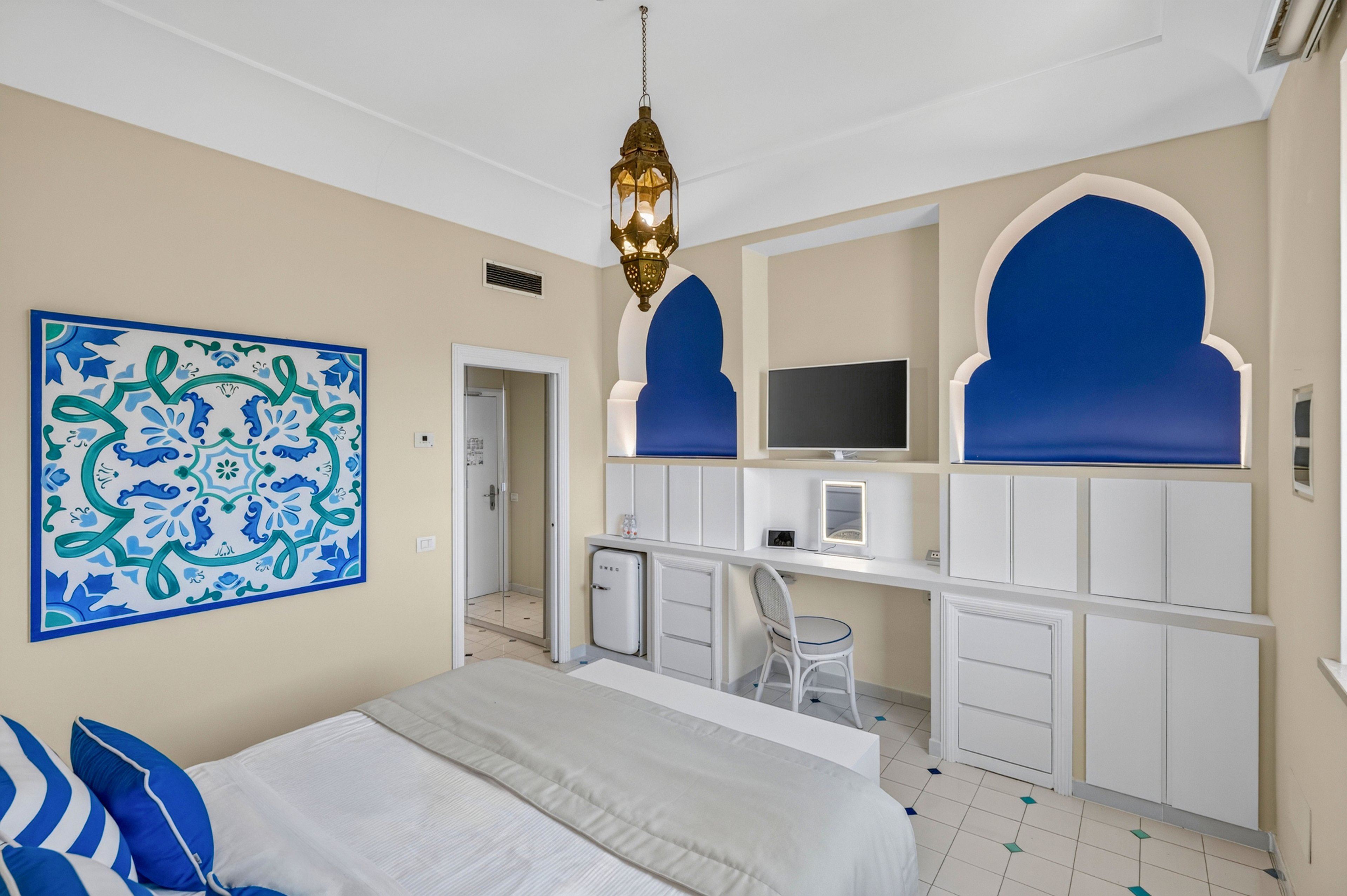 A light-filled hotel bedroom features a neatly made bed with blue and white accent pillows. The room includes a built-in white desk and storage units, with a wall-mounted television above and a small refrigerator to the side. Decorative arched niches with deep blue interiors and a patterned blue artwork add a Mediterranean-inspired touch. A brass hanging lantern and tiled floor complete the clean, modern coastal aesthetic.