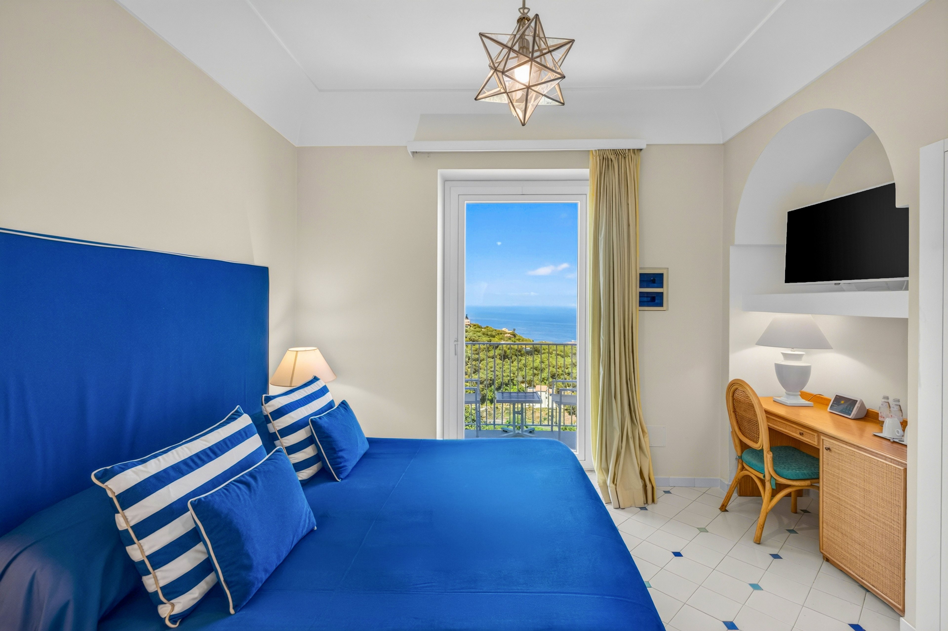 A neatly arranged hotel room features a large blue bed with striped pillows as the focal point. The space includes a wooden desk with a chair, table lamp, and wall-mounted television on the right side. A glass door opens to a balcony overlooking a lush green hillside and distant blue sea under a clear sky. Light-colored walls, tiled floor, and a star-shaped ceiling light create a fresh, Mediterranean-inspired atmosphere.