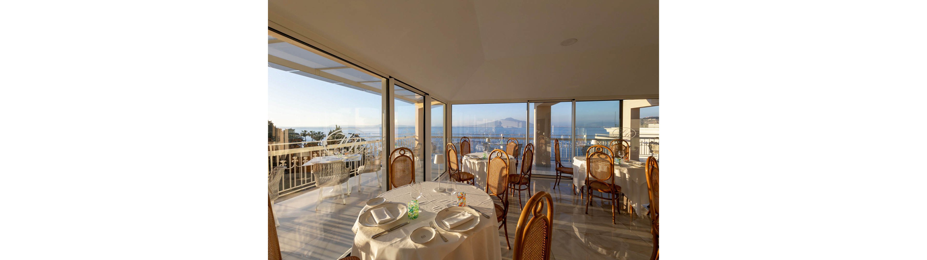 Elegant round tables are set with white tablecloths and plates inside a bright seaside restaurant. Large floor-to-ceiling windows reveal a panoramic view of the ocean, coastline, and distant mountains at sunset. Warm natural light fills the space, highlighting wicker-backed chairs and neatly arranged table settings. The atmosphere feels calm and inviting, ideal for relaxed dining with scenic waterfront vistas.