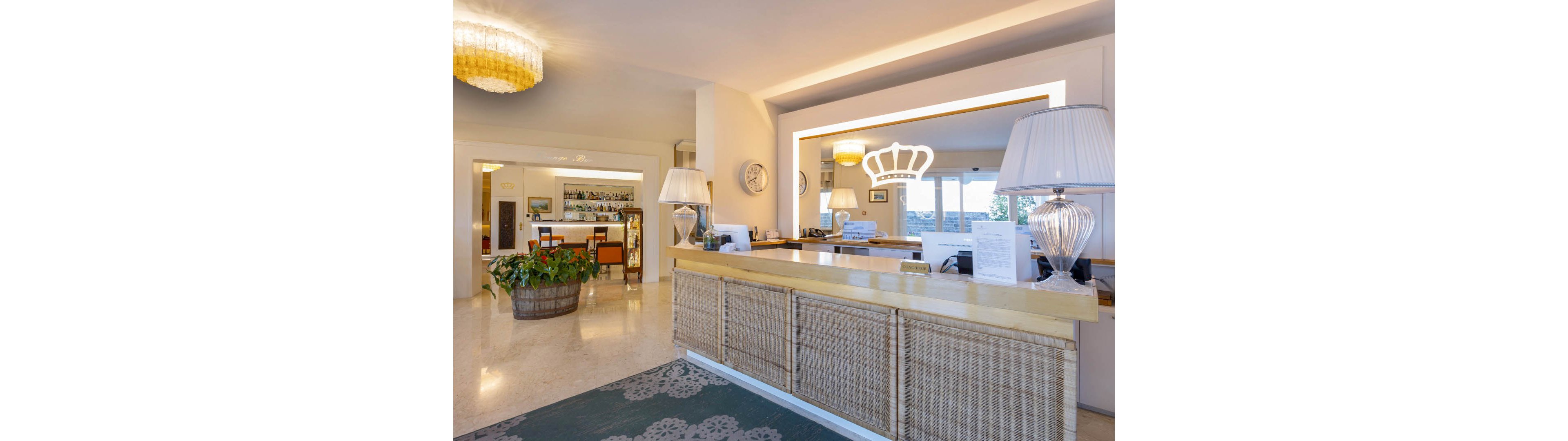 The image shows a bright and welcoming hotel reception desk with a woven front panel and light wooden countertop. A large illuminated crown-shaped wall feature frames the reception area, while table lamps and decorative plants add warmth. In the background, a bar with bottles and glassware is visible, along with seating and large windows letting in natural light. The polished floor and soft lighting create an inviting, contemporary atmosphere suitable for guests checking in or relaxing.