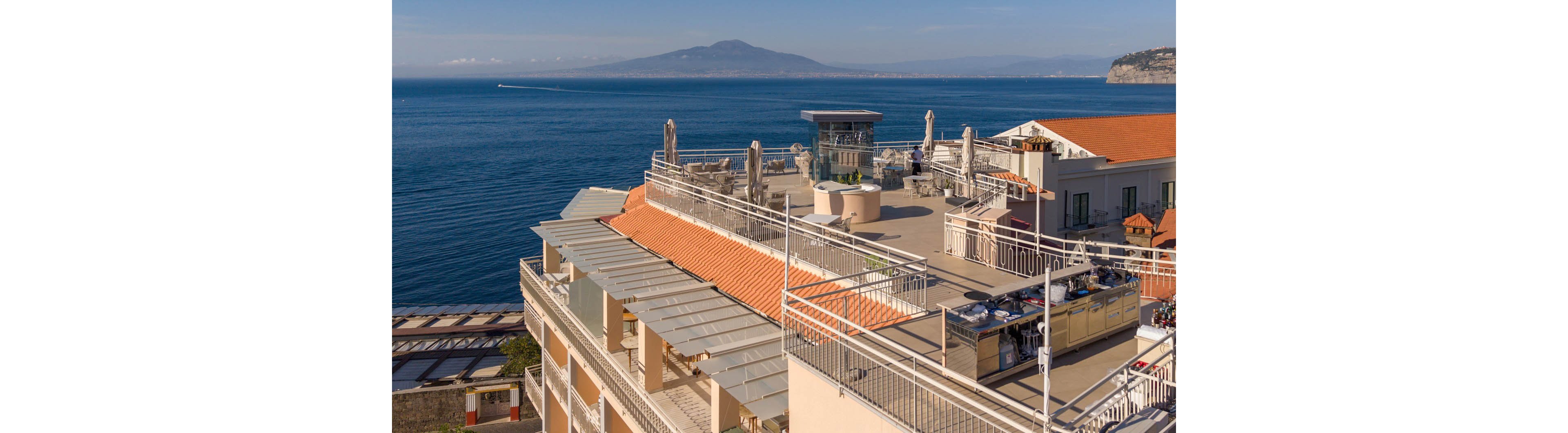 A sunlit rooftop terrace of a coastal building overlooks a calm blue sea and distant mountains. The terrace features railings, shaded areas, and outdoor seating under clear skies. Warm tones of the roof contrast with the deep blue water, creating a serene Mediterranean atmosphere. A boat is visible in the distance, emphasizing the seaside setting.