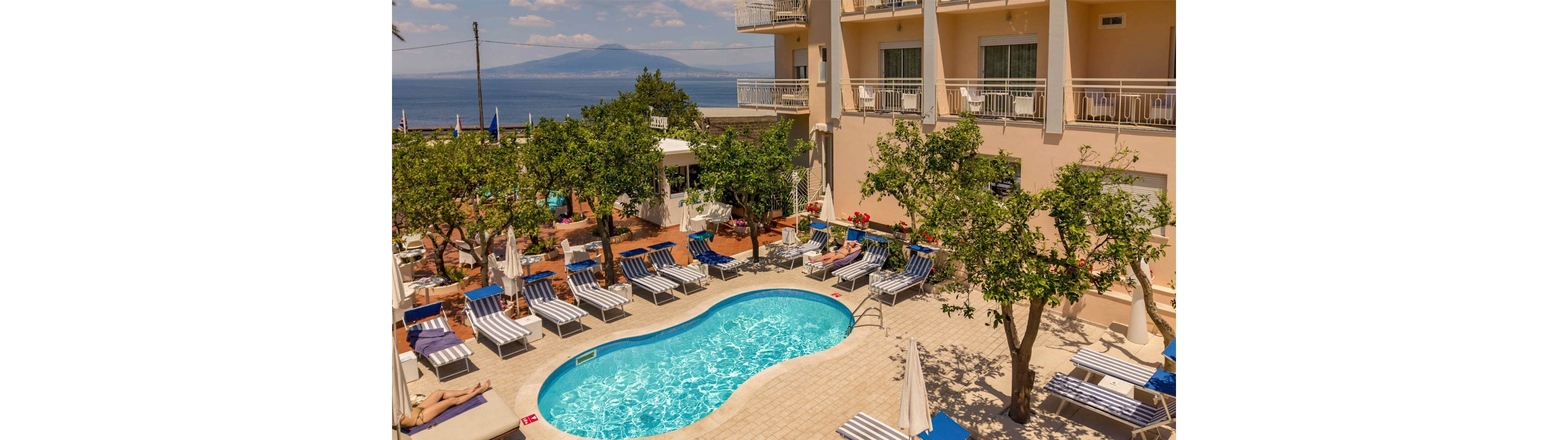 An outdoor hotel pool area is surrounded by striped lounge chairs and umbrellas under a clear, sunny sky. Guests relax on sunbeds beside the small, curved swimming pool set on a tiled terrace. Green trees provide partial shade, while the multi-story hotel building with balconies rises on one side. In the background, a calm sea and distant mountains complete the scenic coastal view.