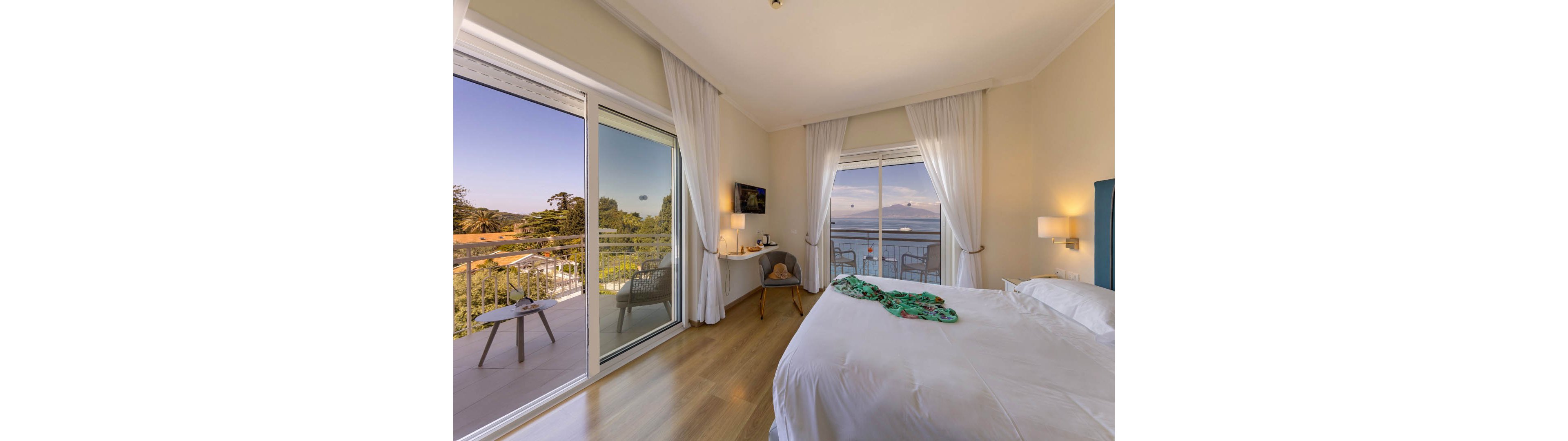 A sunlit hotel bedroom features a neatly made bed with white linens and a colorful garment laid across it. Large sliding glass doors open to a balcony with outdoor chairs overlooking a green landscape and distant hills. Inside, a small desk with a chair, wall-mounted television, and soft lighting create a comfortable, modern atmosphere. Floor-to-ceiling curtains, light walls, and wooden flooring contribute to the airy, relaxing feel of the room.