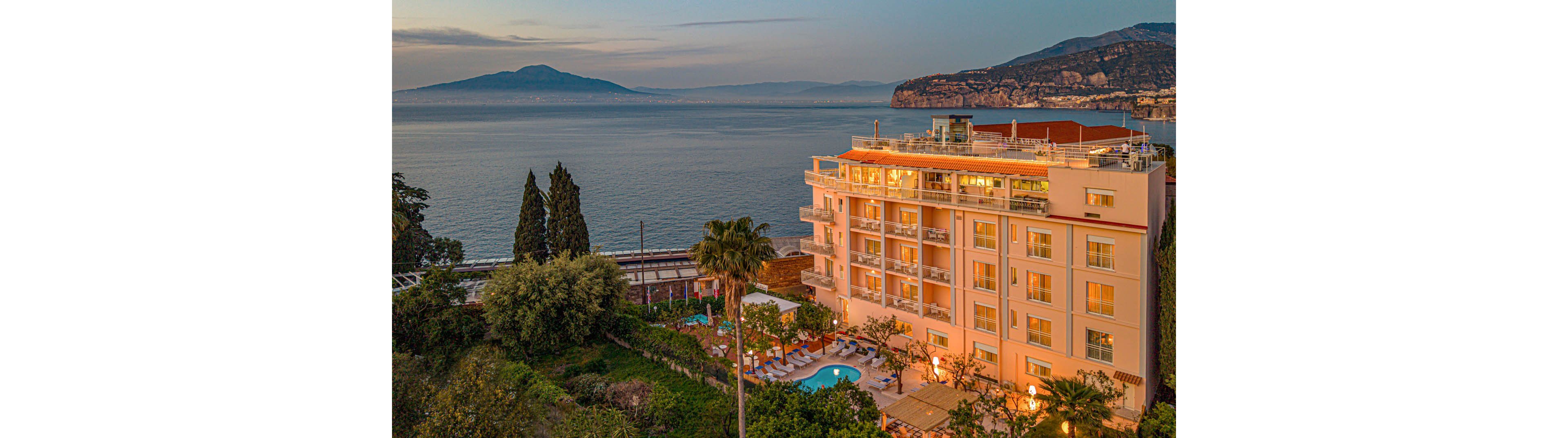 A multi-story seaside hotel stands above a lush garden and pool area, facing a calm bay at sunset. Warm lights from the building and surrounding pathways contrast with the soft blue and orange tones of the sky and water. Tall trees and greenery frame the foreground, while distant hills and a large island or peninsula rise across the bay. The overall scene suggests a tranquil vacation destination with scenic ocean views.