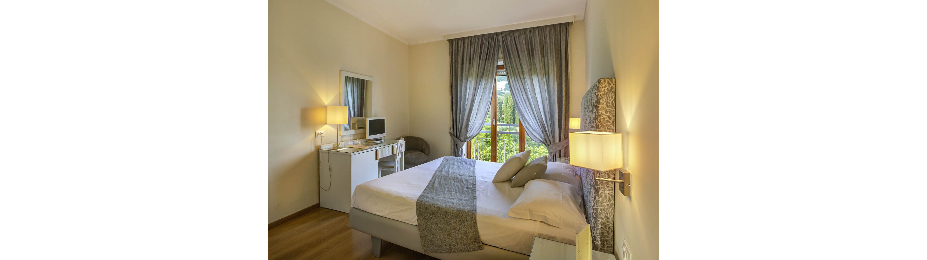Bright, neatly arranged bedroom featuring a double bed with neutral bedding and a patterned headboard. A small desk with a computer monitor and mirror sits against the wall, accompanied by a white chair and table lamp. Floor-to-ceiling curtains frame a large window that opens to greenery outside, bringing natural light into the room. The overall atmosphere is calm and inviting, with soft lighting and warm wooden flooring.
