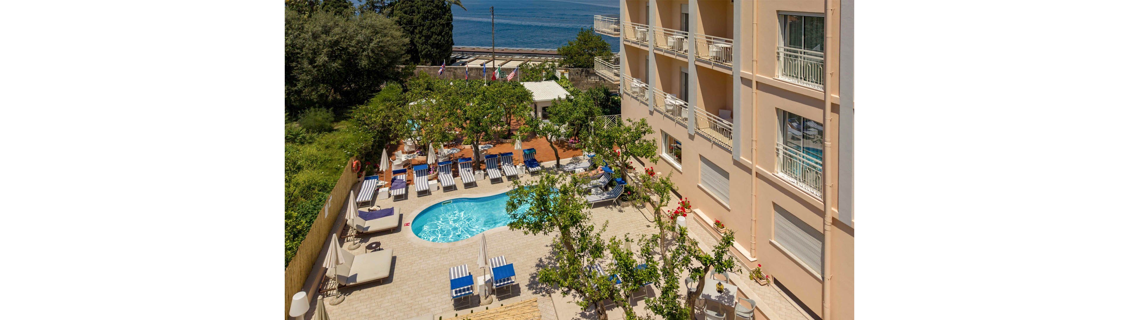 An elevated view shows a hotel courtyard featuring a small curved swimming pool surrounded by lounge chairs and umbrellas. The light-colored building with multiple balconies frames the scene on one side, while trees and greenery add shade around the deck area. Beyond the courtyard, a coastal road and the blue sea are visible in the background, suggesting a seaside location. The overall atmosphere is sunny, relaxed, and inviting for vacation guests.