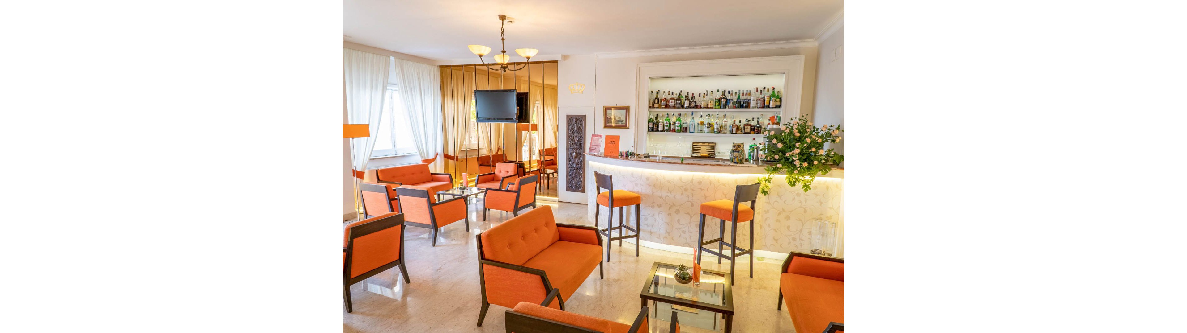 A cozy hotel lounge bar features multiple orange armchairs and stools arranged around glass-topped tables. The bar counter is illuminated with warm lighting and displays a wide selection of bottles on white shelves behind it. Large windows with sheer white curtains and golden drapes brighten the room with natural light. The overall atmosphere is inviting and modern, suitable for socializing or relaxing with a drink.