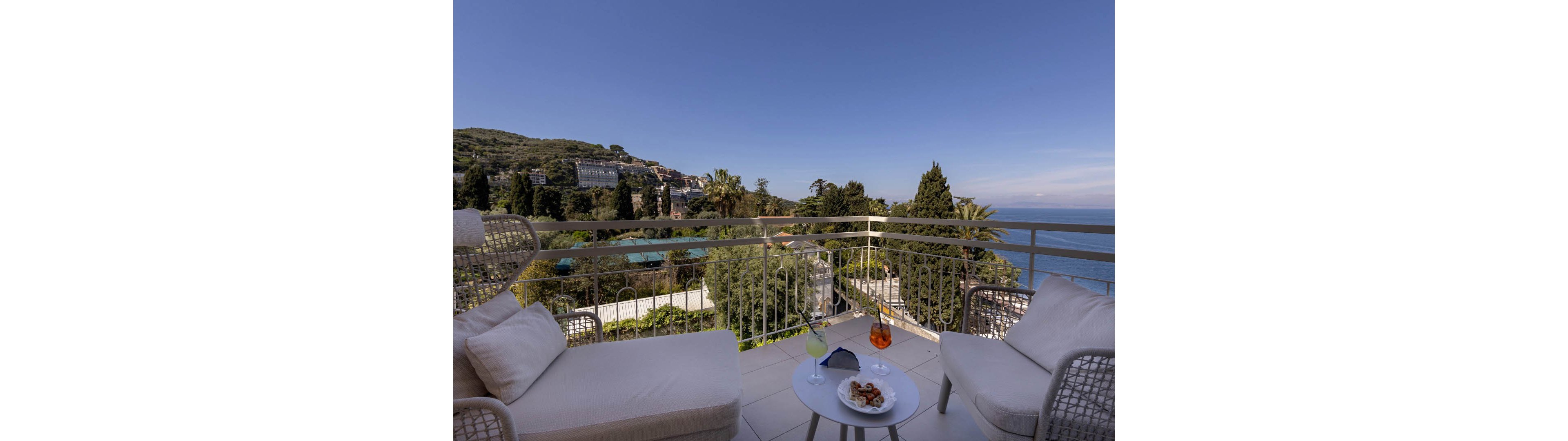 A cozy terrace seating area overlooks a lush coastal landscape and the sea under a clear blue sky. Two cushioned lounge chairs surround a small round table holding colorful cocktails and a plate of snacks. The balcony railing frames distant hills, trees, and buildings, creating a relaxed vacation atmosphere. The scene suggests leisure, comfort, and outdoor enjoyment in a scenic seaside setting.