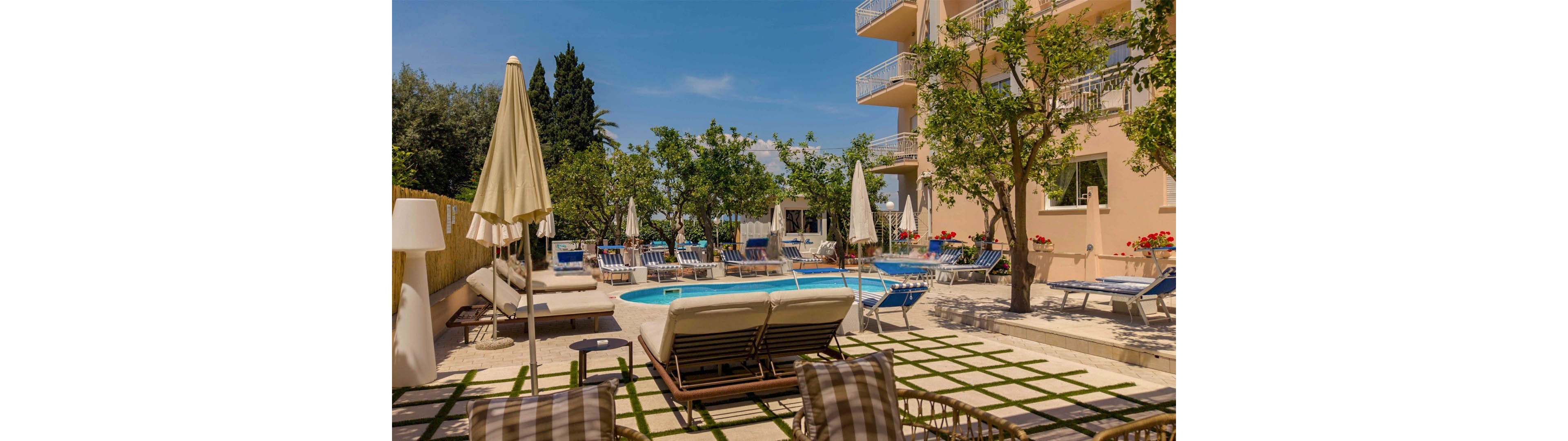 An inviting hotel courtyard features a small outdoor swimming pool surrounded by sun loungers and umbrellas. Trees and greenery provide shade and a relaxed atmosphere around the pool area. A multi-story building with balconies overlooks the courtyard, suggesting guest rooms with pool views. The bright daylight and clear blue sky create a warm, summery vacation setting.