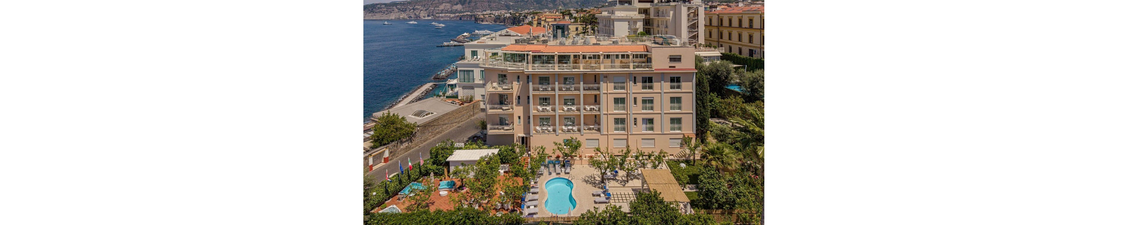 An elegant multi-story hotel stands along a rocky coastline overlooking the sea. The building features numerous balconies and a rooftop terrace with panoramic views. In the foreground, a curved swimming pool is surrounded by sun loungers, greenery, and landscaped gardens. Nearby structures and neighboring hotels indicate a coastal town setting with a relaxed vacation atmosphere.