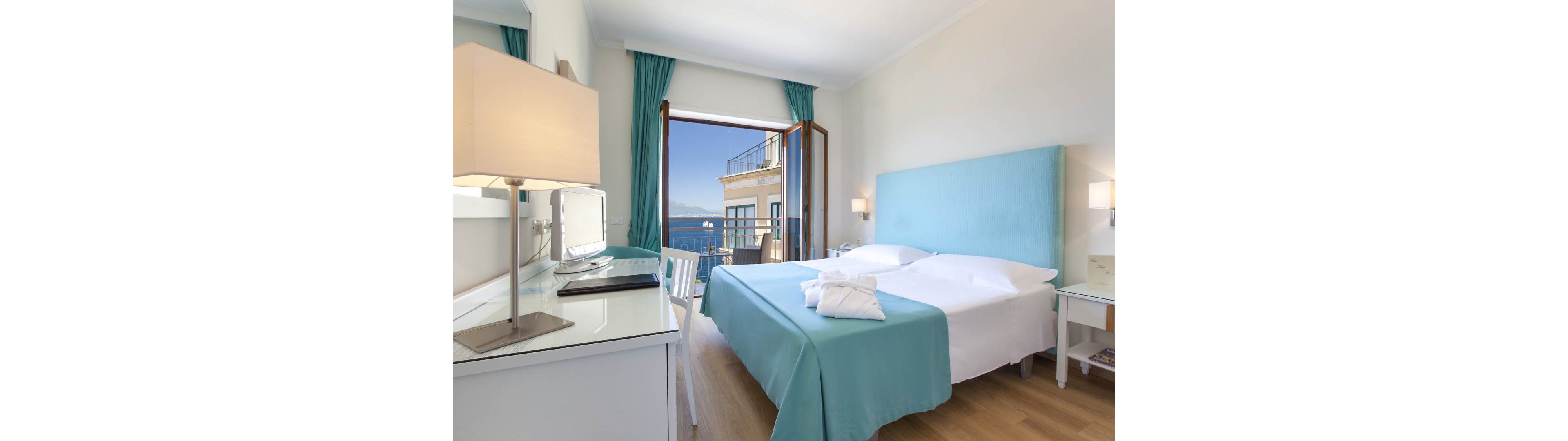 A modern hotel room features a neatly made double bed with turquoise bedding and a matching upholstered headboard. Large glass doors open onto a balcony, revealing a scenic ocean view and nearby building. A desk with a lamp, chair, and television sits opposite the bed, while bedside tables hold additional lamps. Natural light fills the space, highlighting the clean, minimalist decor and light wood flooring.