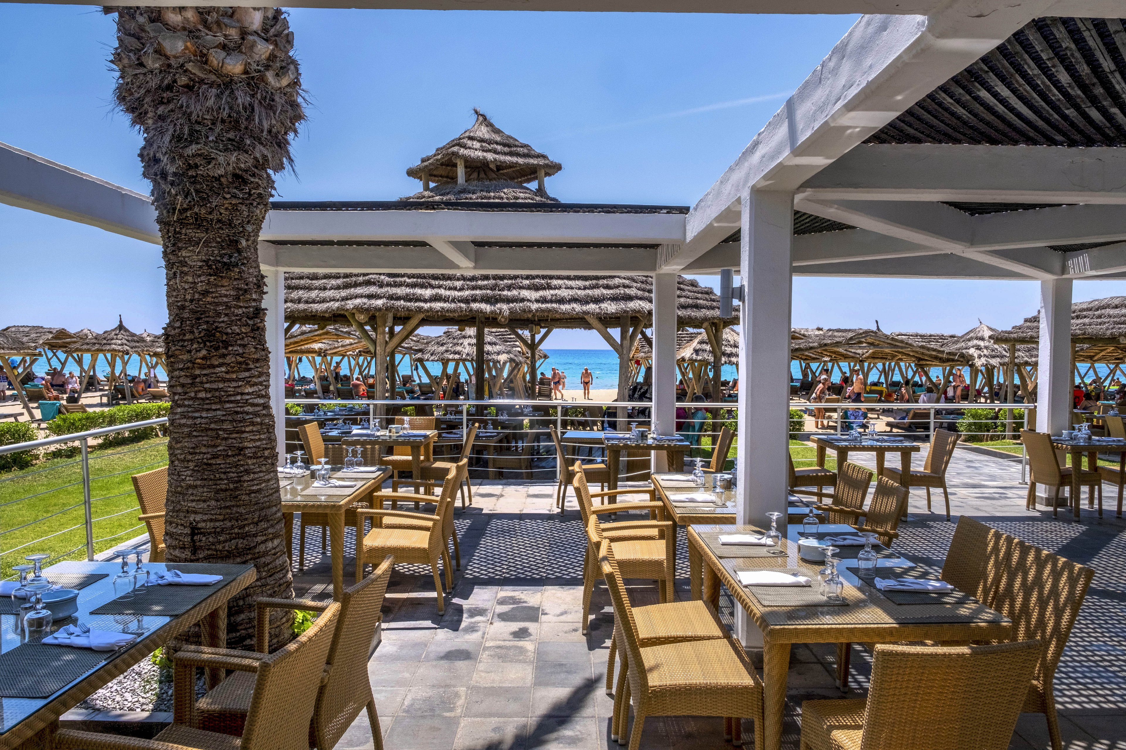 An open-air restaurant terrace overlooks a sunny beach lined with thatched umbrellas. Neatly arranged tables and wicker chairs sit under a shaded pergola, set with plates, glasses, and cutlery. Palm trees and green lawn separate the dining area from the sandy shore and blue ocean. A few people can be seen walking near the water, adding a relaxed holiday atmosphere.