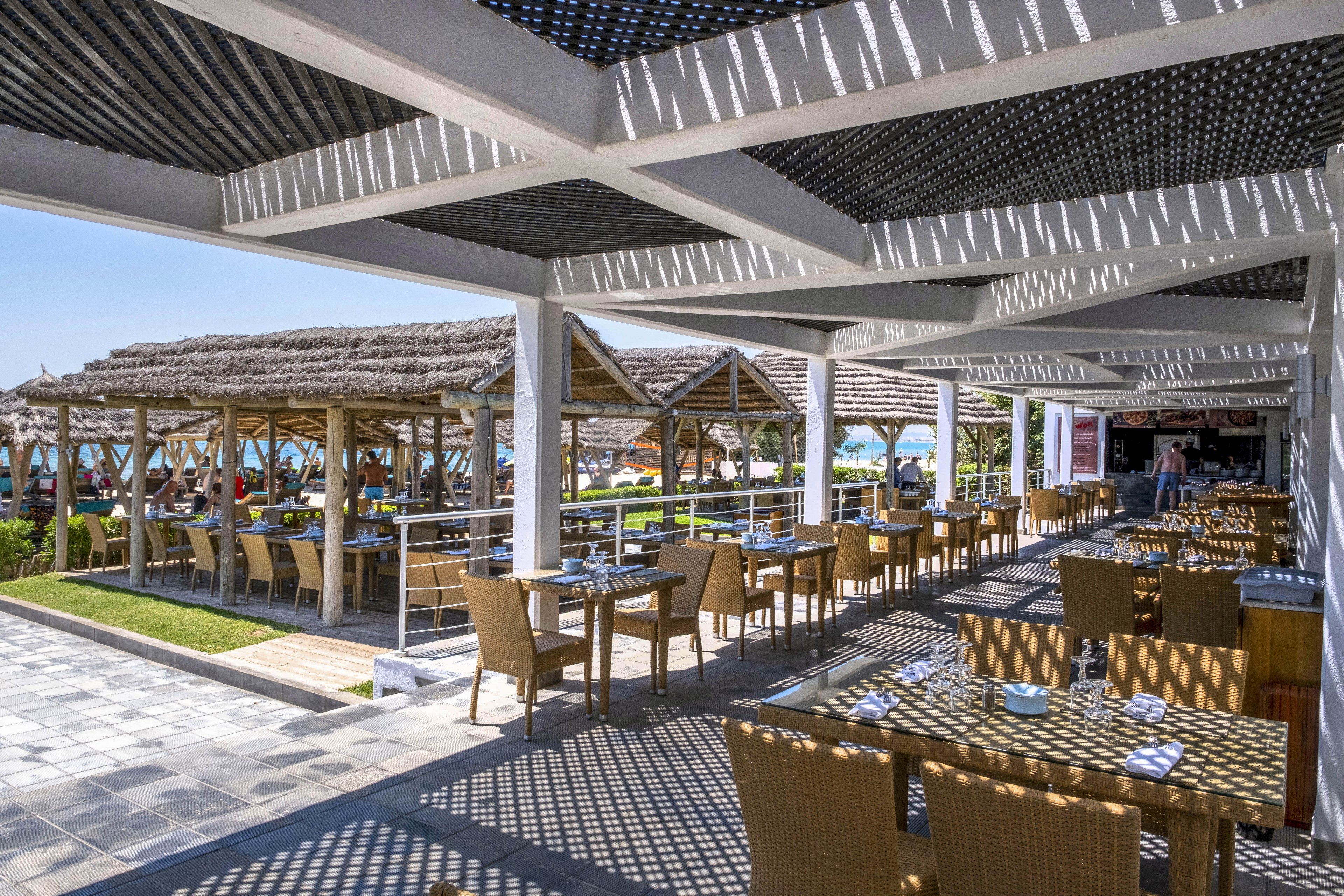 An open-air restaurant with wicker chairs and wooden tables is set under a modern pergola casting patterned shadows. The dining area overlooks a row of thatched-roof cabanas and a bright blue sea in the background. Place settings with plates, glasses, and napkins are arranged neatly on the tables, suggesting readiness for guests. The sunny coastal setting and clear sky create a relaxed, vacation-like atmosphere.