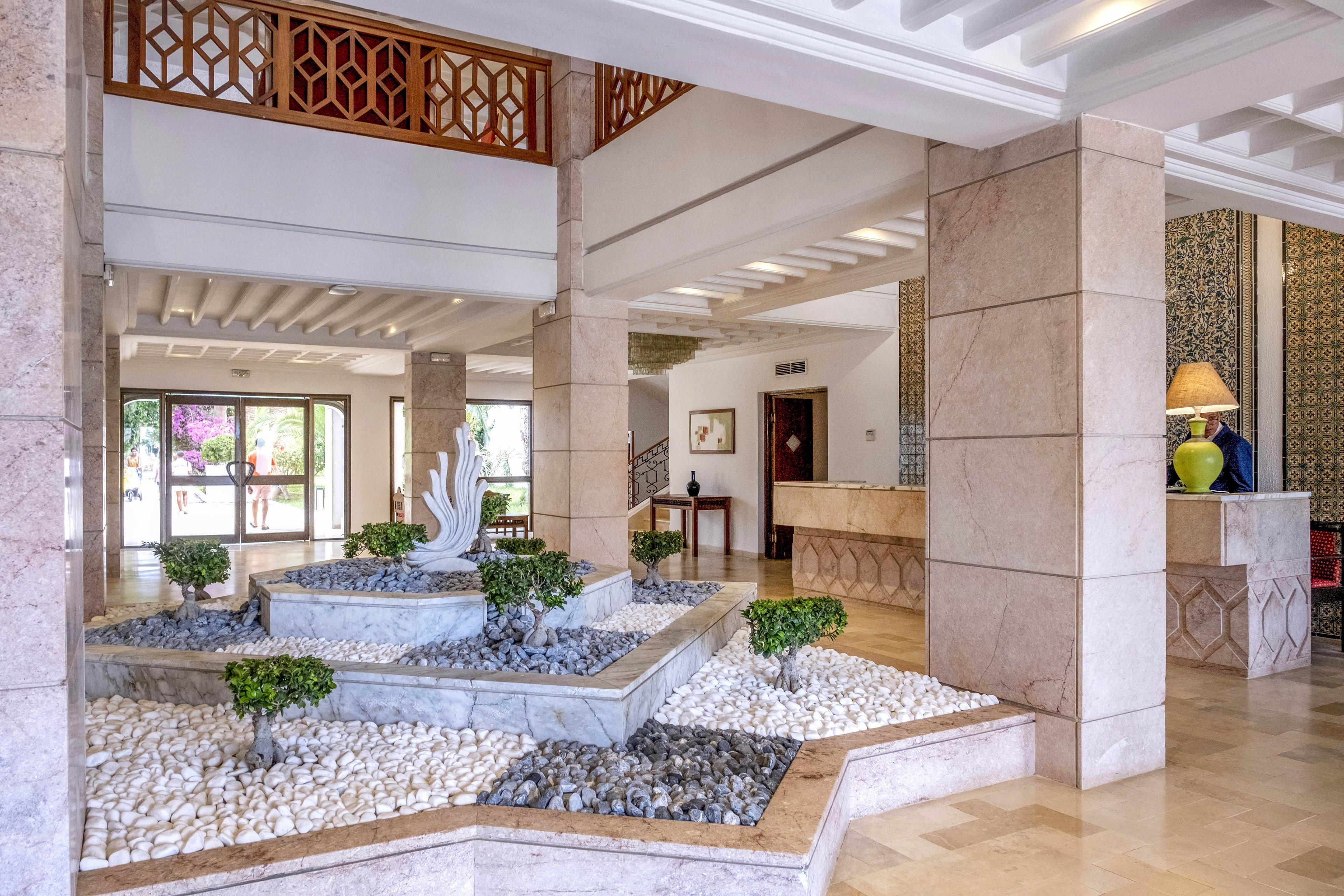 A spacious hotel lobby features a central indoor stone garden with layered platforms and a sculptural centerpiece. White and gray pebbles surround neatly trimmed small shrubs, adding a natural accent to the polished floor. The reception desk and seating area are visible in the background, framed by patterned wall panels and warm lighting. Large glass doors at the far end let in daylight and a view of colorful flowers outside.