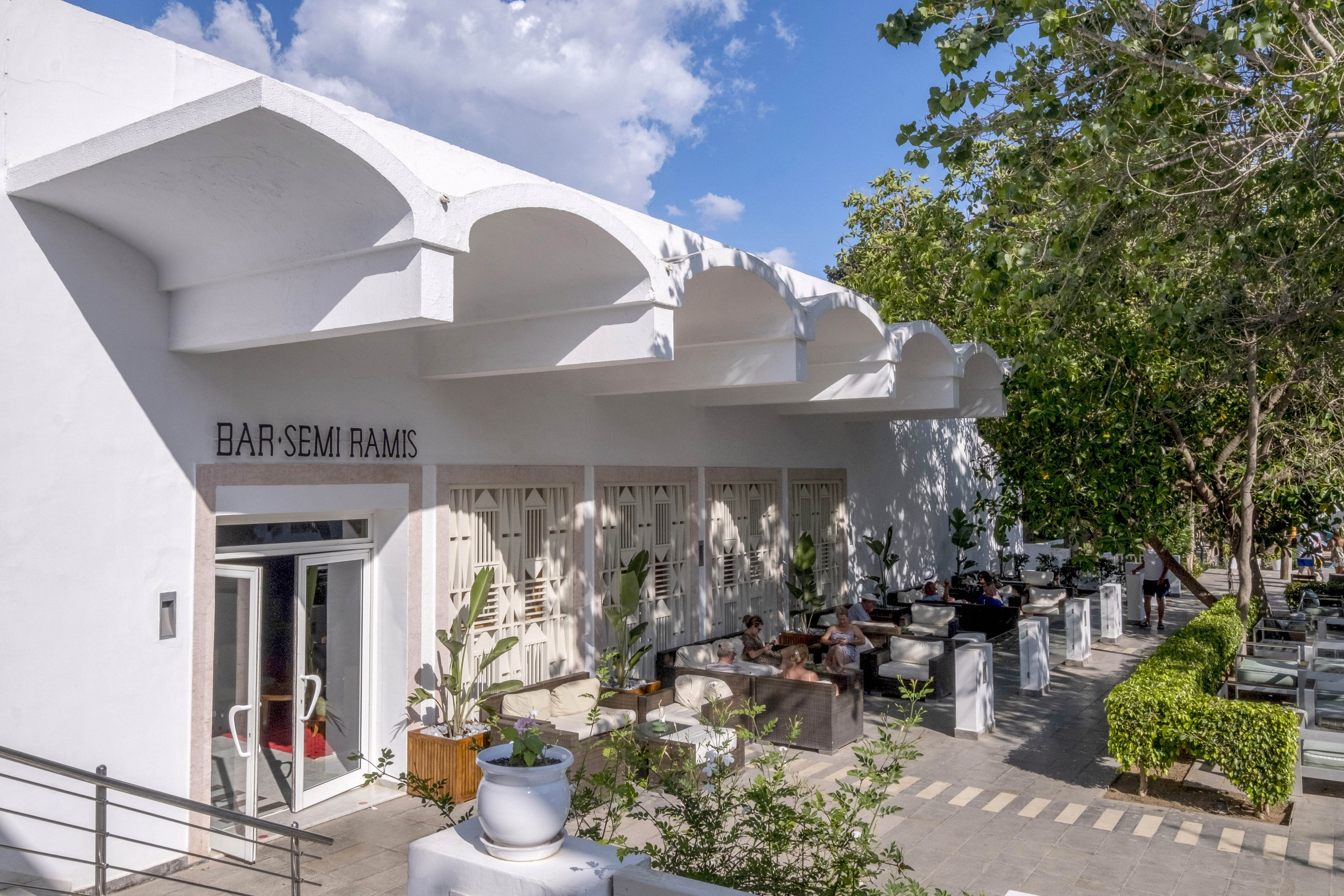 A modern white building houses a bar with an outdoor terrace. The sign above the entrance reads "BAR SEMI RAMIS" in clear letters. Guests sit at tables under the shade of trees and architectural overhangs. Potted plants, hedges, and contemporary furniture create a relaxed, stylish atmosphere on a sunny day.