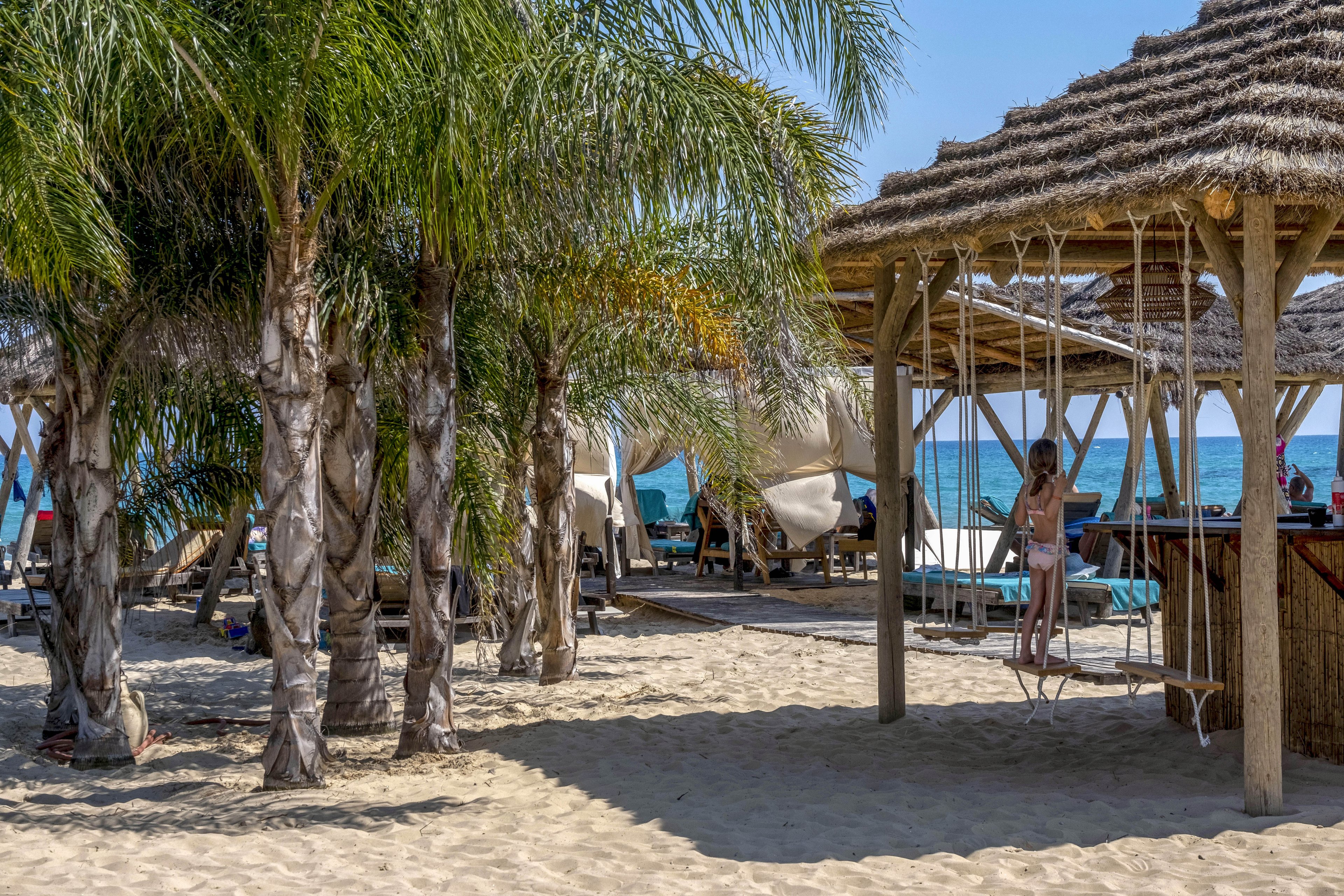 A sandy beach is lined with tall palm trees and rustic wooden cabanas. A woman in swimwear stands on a swing under a thatched-roof structure near a beach bar. Lounge chairs and shaded daybeds face the calm blue sea in the background. The scene suggests a relaxed, tropical vacation atmosphere on a sunny day.