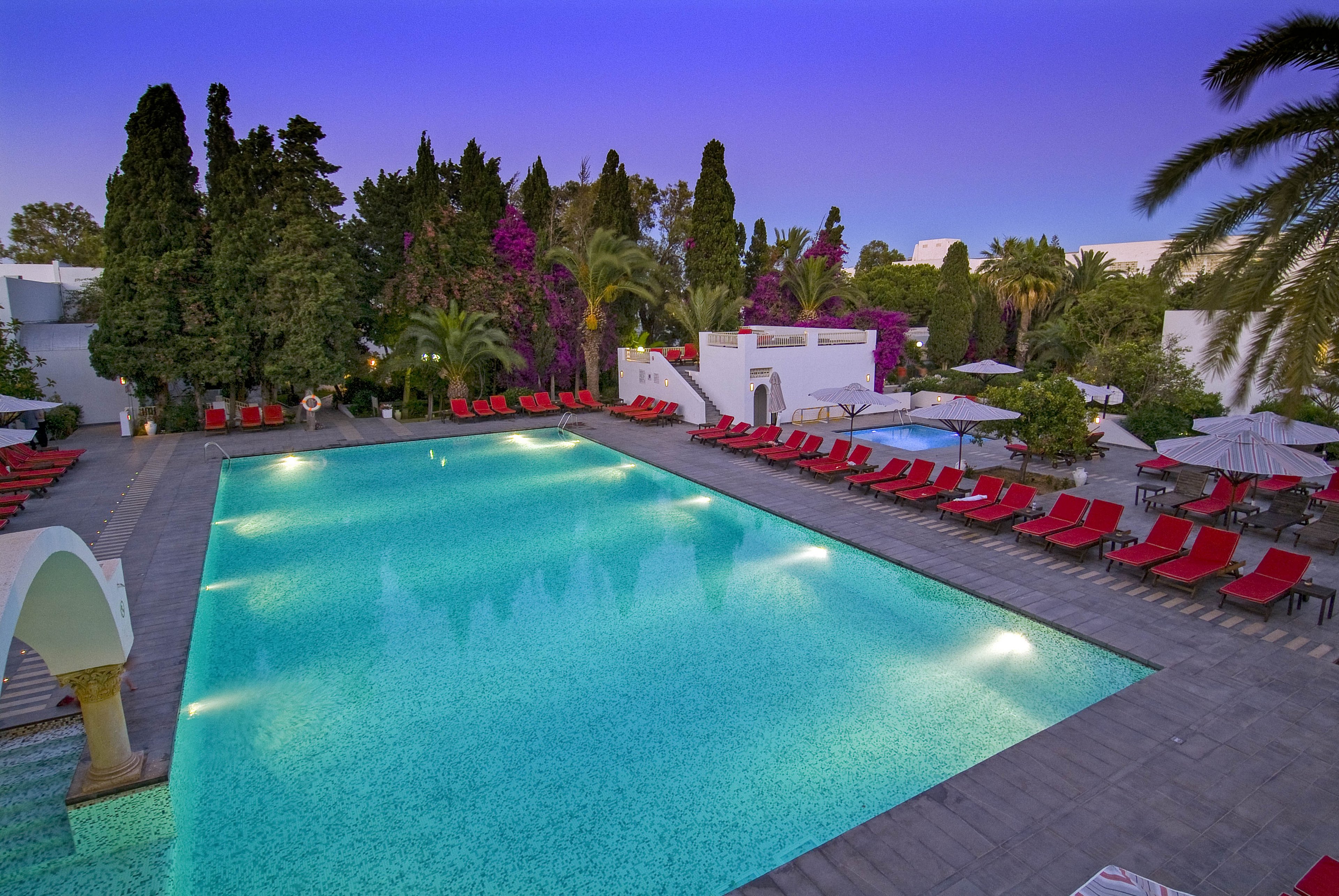 A large illuminated swimming pool is surrounded by red lounge chairs and umbrellas at a resort. Tall trees, palms, and flowering bushes frame the pool area, creating a lush garden atmosphere. The sky transitions from deep blue to purple, suggesting early evening or twilight. White buildings with arches and terraces add a Mediterranean or tropical feel to the tranquil scene.