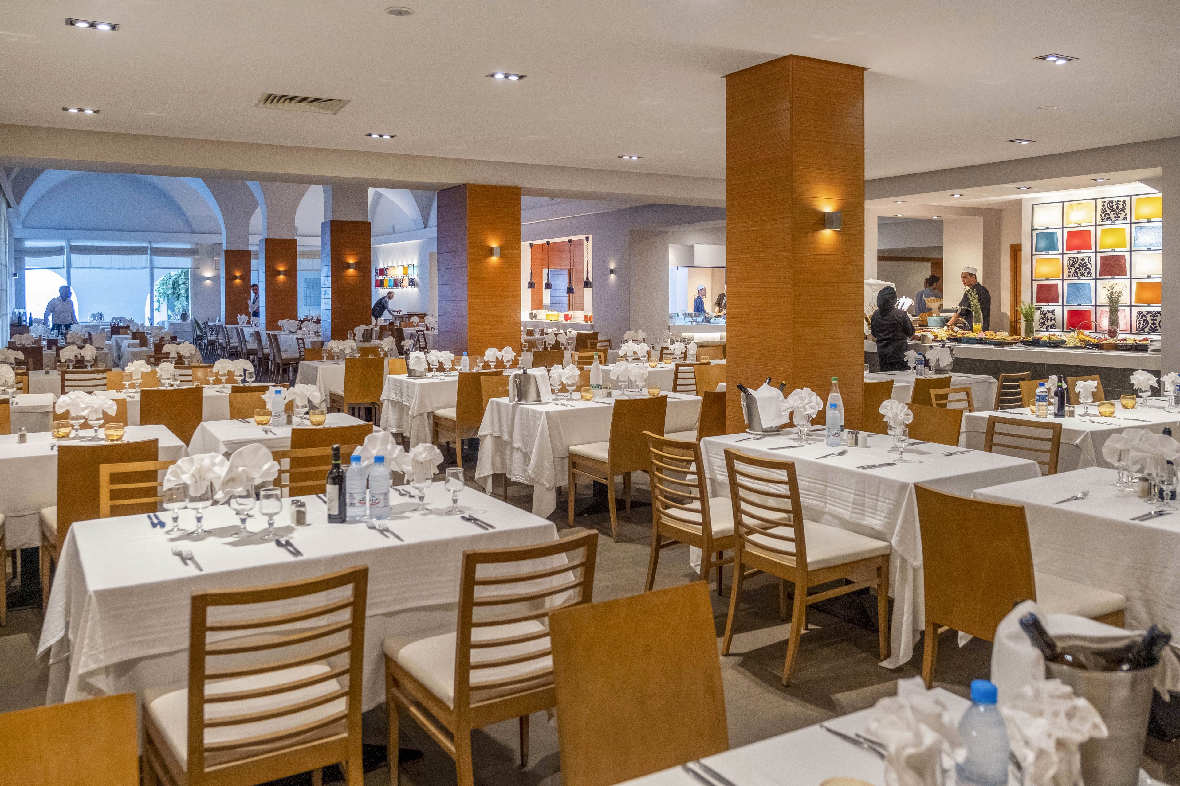 A spacious restaurant dining room is arranged with rows of neatly set tables covered in white tablecloths. Wooden chairs and warm wood-paneled columns create a modern yet inviting atmosphere. Napkins are folded decoratively, and glassware and cutlery are carefully placed, suggesting preparation for a meal service. In the background, staff move near a brightly lit bar or buffet area with colorful wall art.