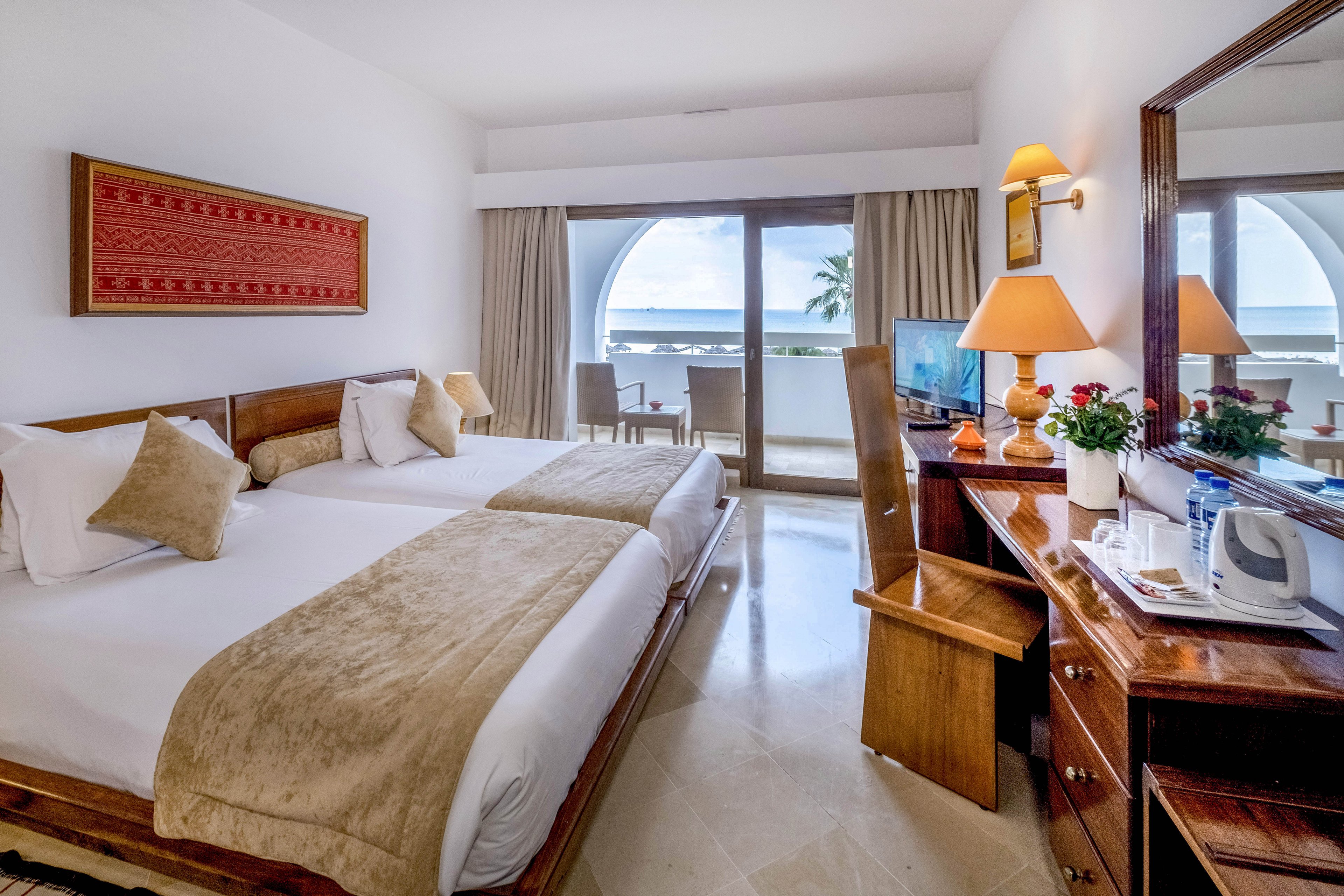 A neatly arranged hotel room features two twin beds with beige throws and multiple pillows. A wooden desk with a chair, table lamp, flowers, and a television sits opposite the beds. Large sliding glass doors open to a balcony with outdoor seating and a partial ocean view framed by white arches. The room is decorated in warm neutral tones, creating a clean and inviting atmosphere.