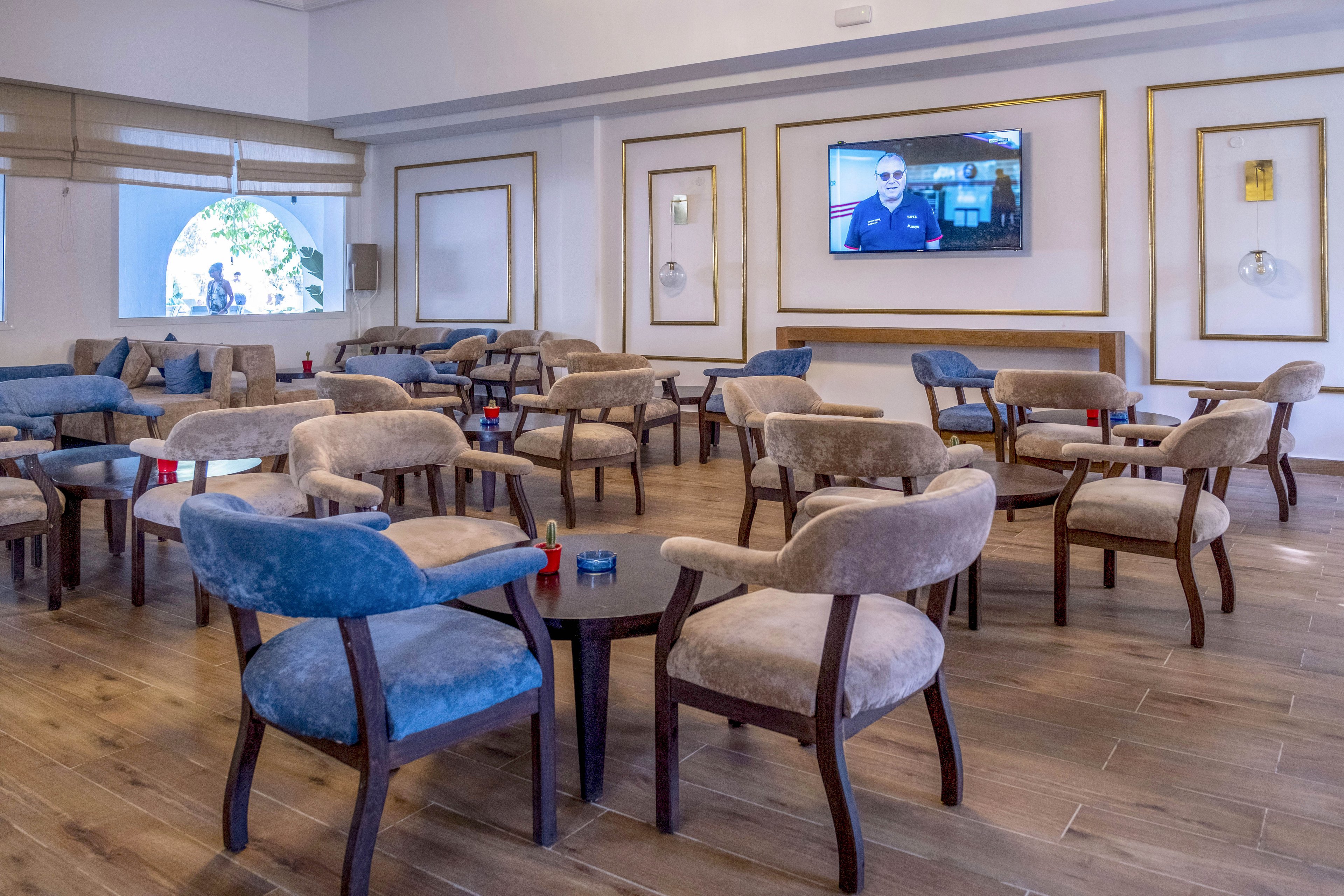 A contemporary lounge area features multiple upholstered chairs and small round tables arranged on a wooden floor. The space appears to be part of a hotel or resort, with neutral-toned decor and framed wall panels. A television mounted on the wall displays a person speaking, adding a casual entertainment element. Large windows and soft lighting create an inviting yet quiet atmosphere, with no guests currently present.
