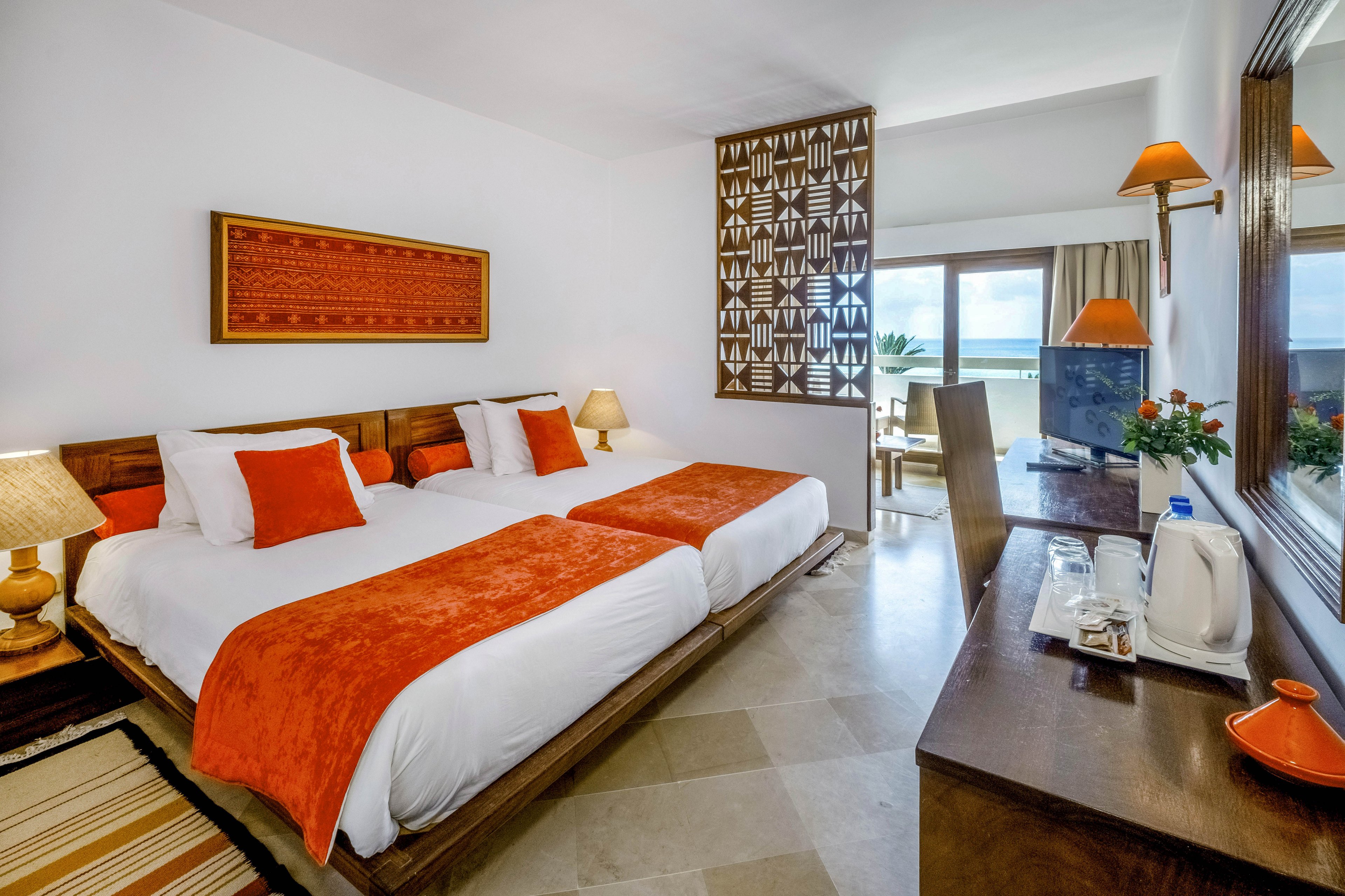 A modern hotel room features two neatly made twin beds with white linens and orange throws and pillows. The space includes a wooden desk with a television, kettle, and refreshments arranged on a tray. Warm lighting from bedside and wall lamps complements the orange and brown decorative accents. A patterned room divider and large window lead to a balcony with outdoor seating and a scenic view.