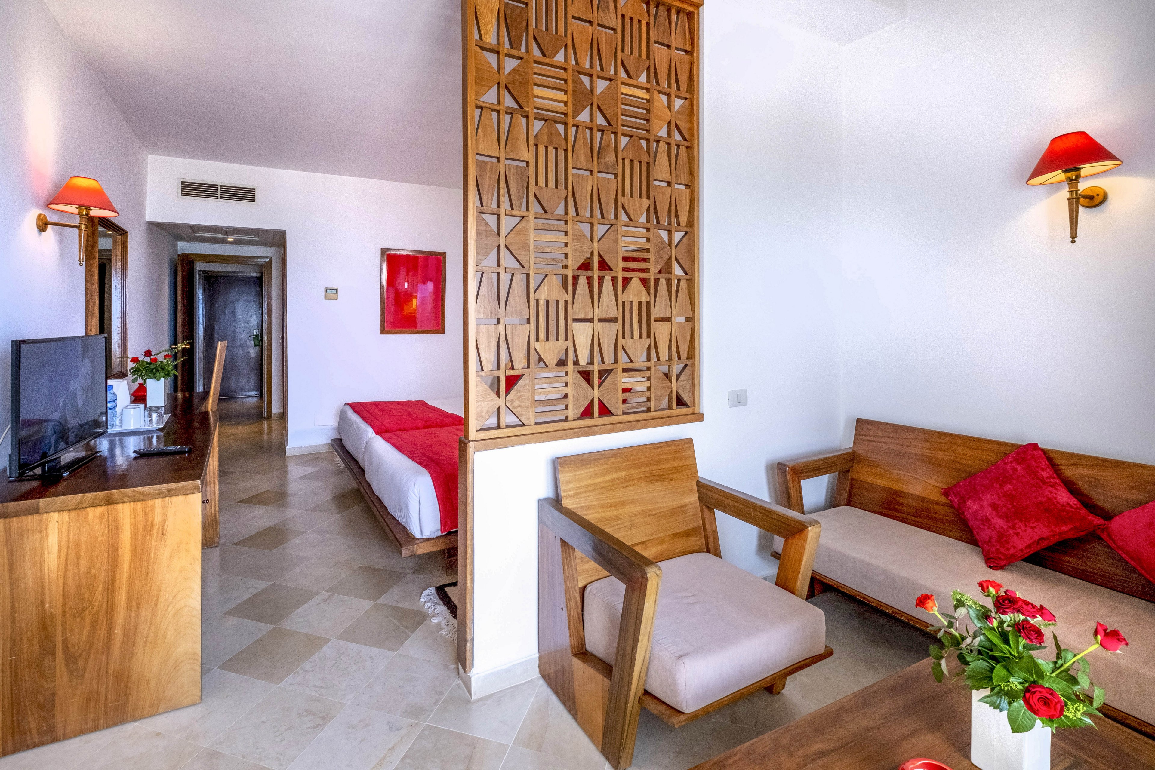 A modern hotel suite features a sitting area with wooden chairs, a sofa, and a small table decorated with red flowers. A decorative wooden partition separates the lounge from the bedroom, where a neatly made bed with a red throw is visible. White walls, tiled floors, and red wall lamps create a clean, minimalist look with warm accents. A television and desk area line one wall, emphasizing comfort and functionality for guests.