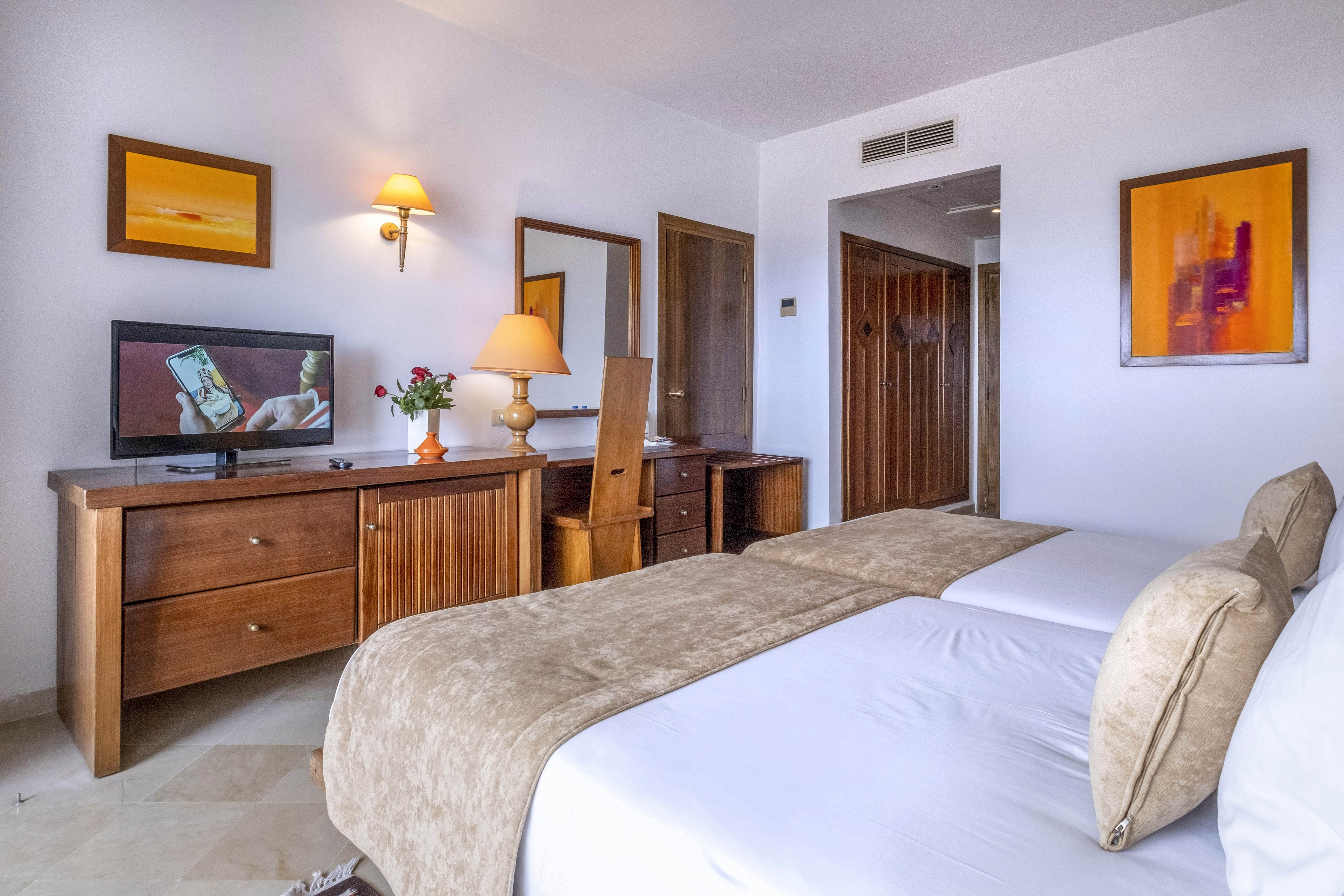 A neatly made twin hotel room features two single beds with beige throws and matching cushions. The space includes a wooden dresser with a flat-screen TV, a desk with a chair, and a table lamp. Warm-toned abstract artwork and a wall sconce add color to the otherwise white walls. The room appears clean, inviting, and well-lit, with a simple contemporary style.