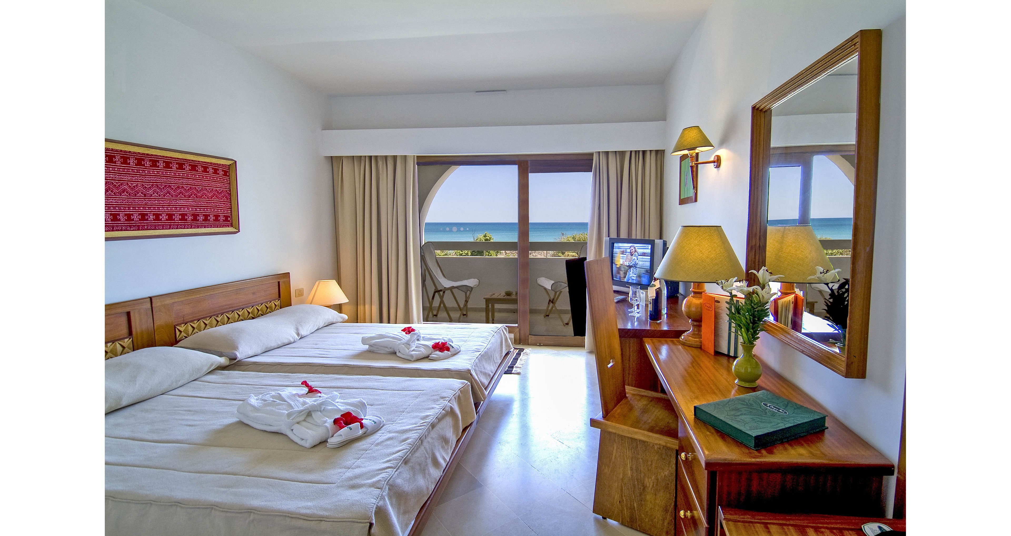 A tidy hotel room features two single beds with folded white robes and red accents placed on top. Wooden furniture, including a desk, mirror, and bedside tables, adds warmth to the bright interior. A sliding glass door opens to a balcony with chairs, offering a clear view of the ocean and blue sky. Soft lighting from table lamps and neutral walls create a calm, inviting atmosphere for guests.