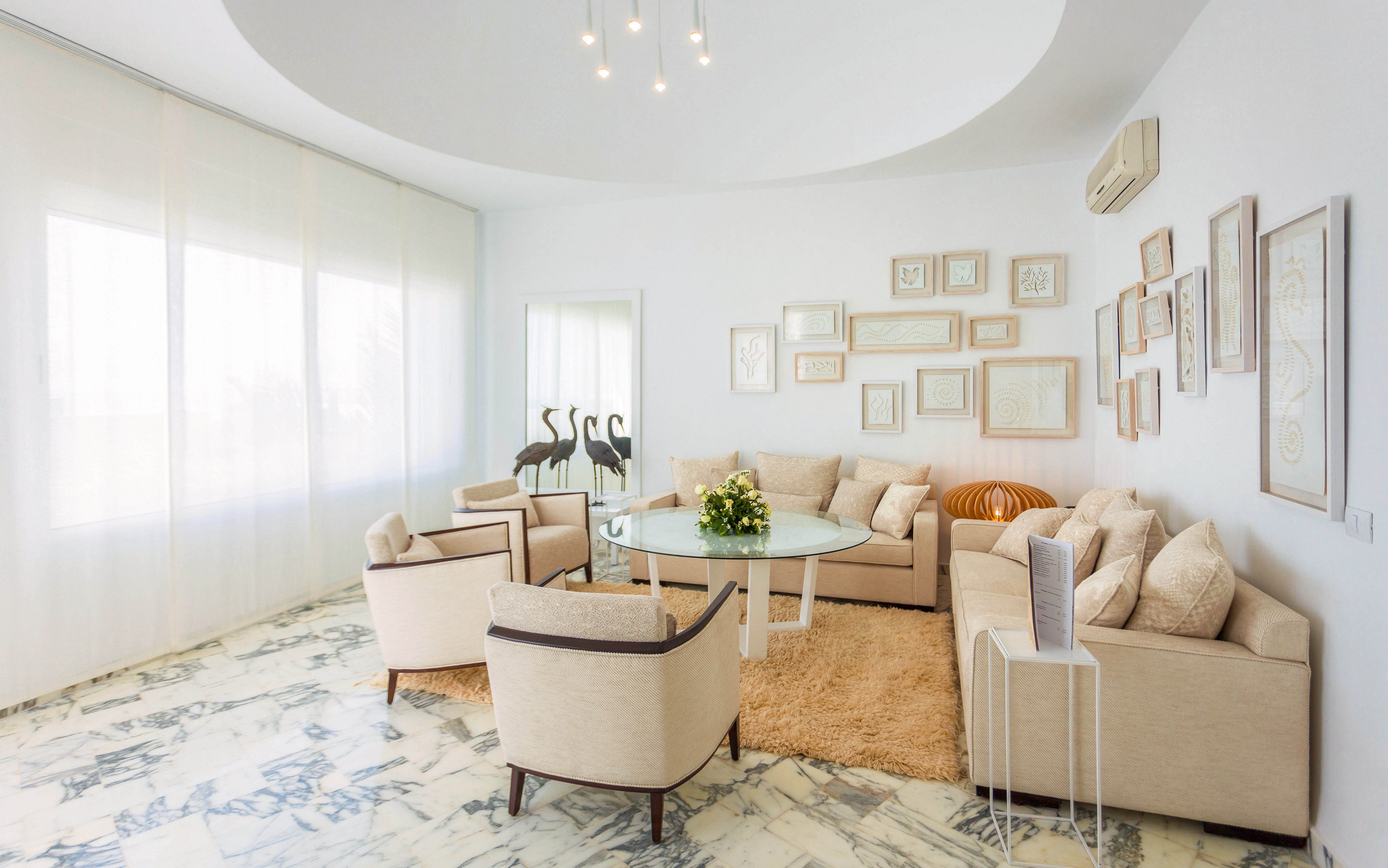 A spacious contemporary living room features cream-colored sofas and armchairs arranged around a round glass coffee table. Large sheer curtains cover tall windows, filling the space with soft natural light. The walls are decorated with an assortment of empty picture frames and minimalist artwork. A shaggy rug, marble-patterned floor, and a bouquet of flowers on the table add texture and warmth to the otherwise light, neutral palette.