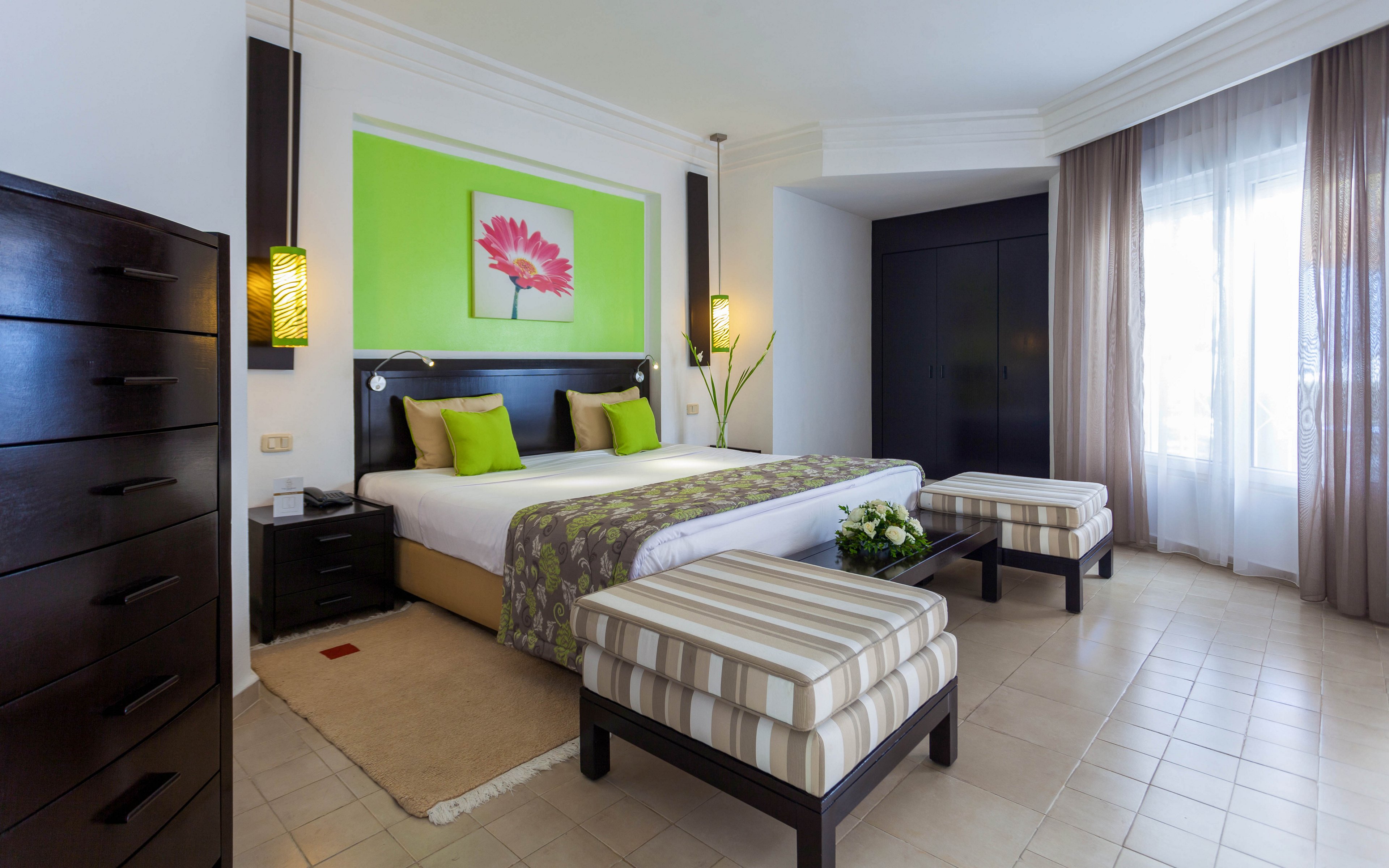A contemporary hotel bedroom features a large double bed with white linens and green accent pillows. The room includes two striped upholstered benches at the foot of the bed and dark wooden furniture. A vivid green accent wall with a floral artwork adds color, complemented by warm pendant lights on either side of the bed. Sheer curtains allow natural light to filter into the clean, minimalist space.