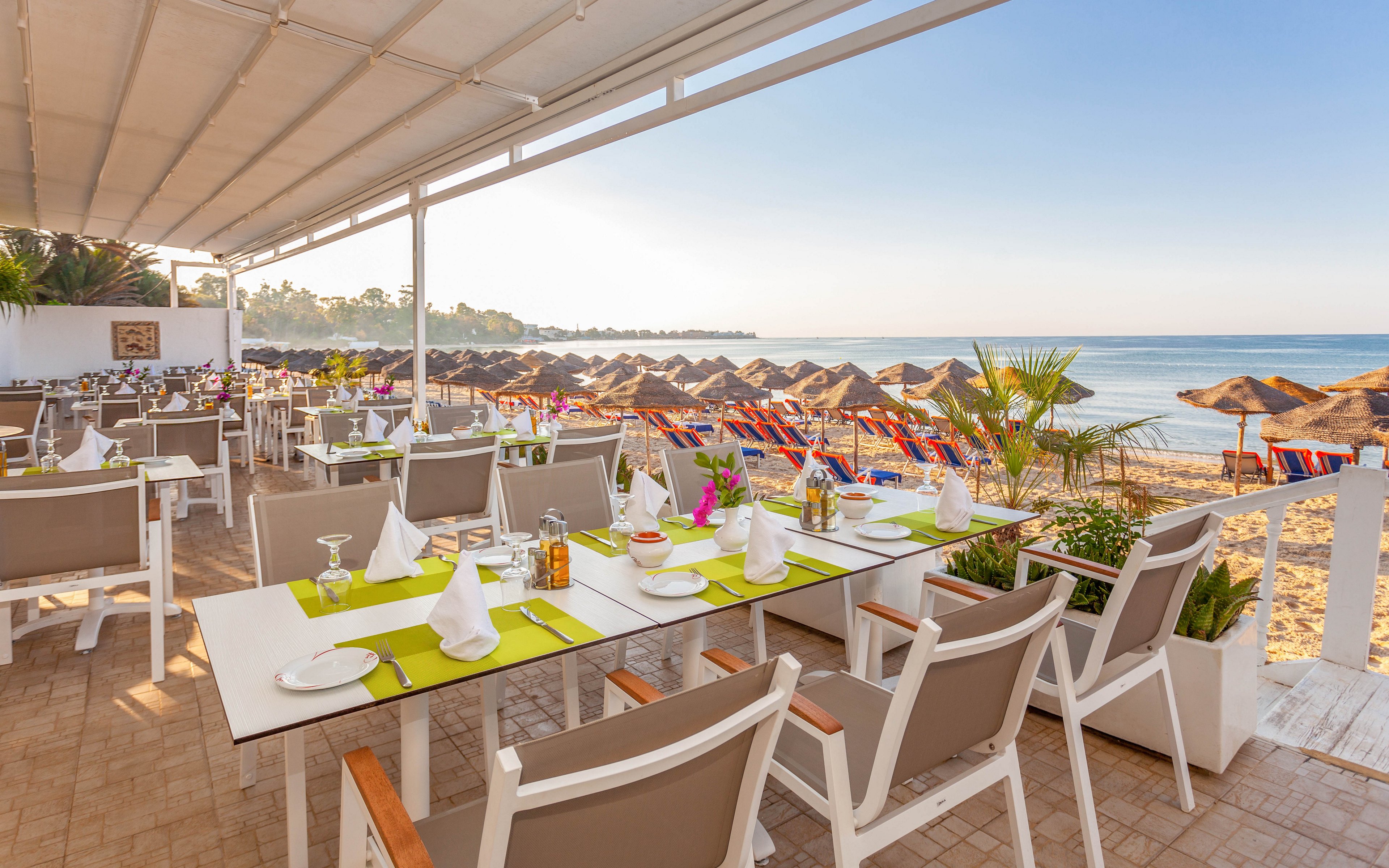 An open-air restaurant terrace is set with neatly arranged tables overlooking a sandy beach. White chairs and tables with green placemats and folded napkins create a clean, modern dining atmosphere. In the background, rows of straw parasols and colorful sun loungers line the shoreline beside calm blue water. The scene is bright and sunny, suggesting a relaxed seaside vacation setting.