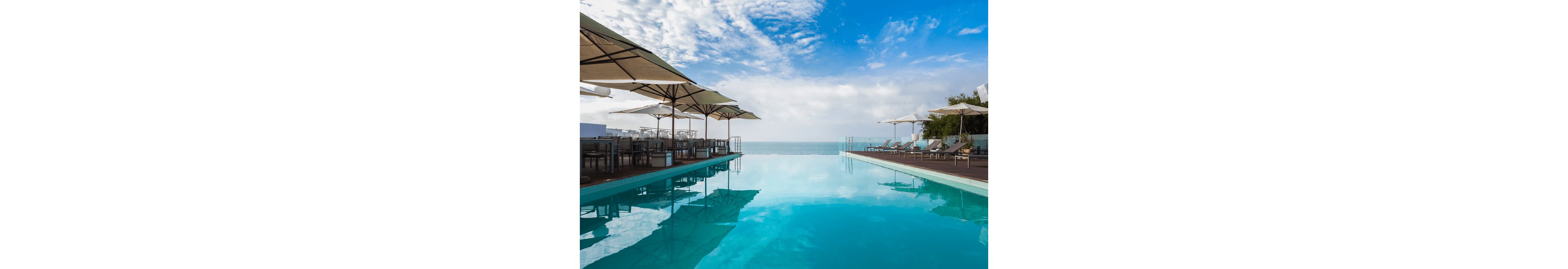A serene infinity pool stretches toward a calm ocean horizon under a bright blue sky with scattered clouds. Sun loungers and large parasols line the wooden deck on both sides of the pool, suggesting a high-end resort or hotel setting. The clear turquoise water reflects the sky and surrounding furniture, enhancing the tranquil atmosphere. The scene evokes relaxation, leisure, and coastal vacation vibes.