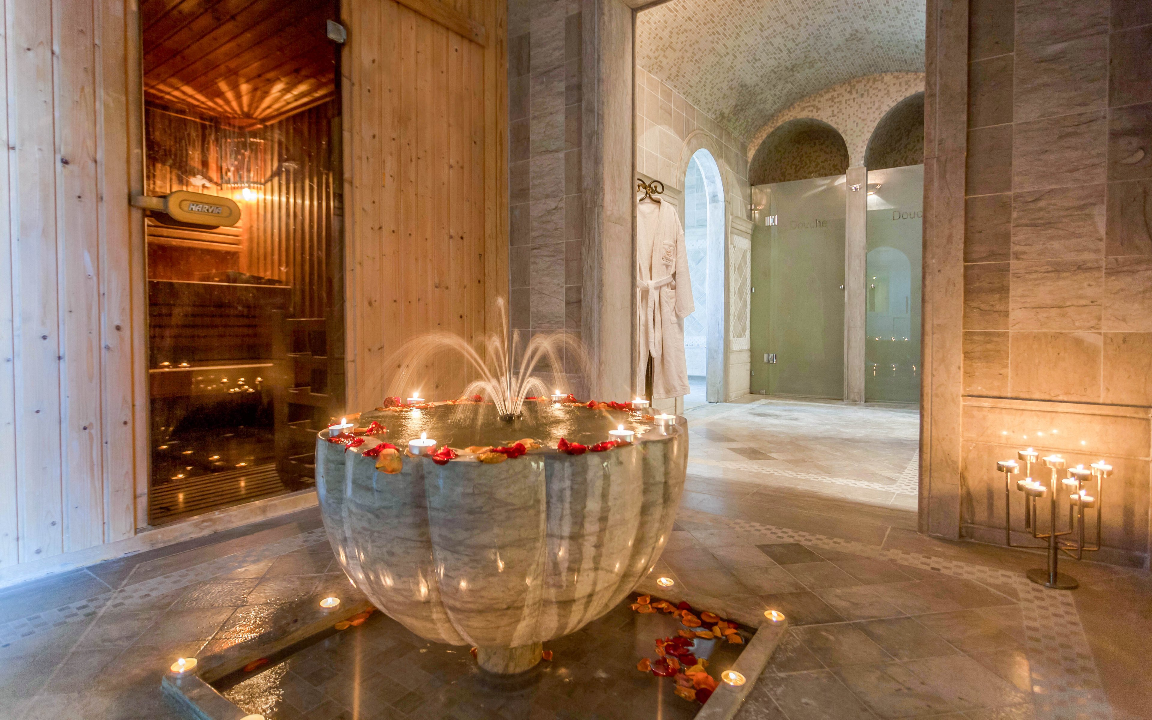 A large stone fountain filled with water and flower petals stands at the center of an elegant spa interior. Small candles surround the fountain, casting a warm glow on the polished stone floor. In the background, a wooden sauna room and arched doorways lead to additional spa areas. Two white bathrobes hang on the wall, enhancing the sense of relaxation and comfort in this tranquil setting.
