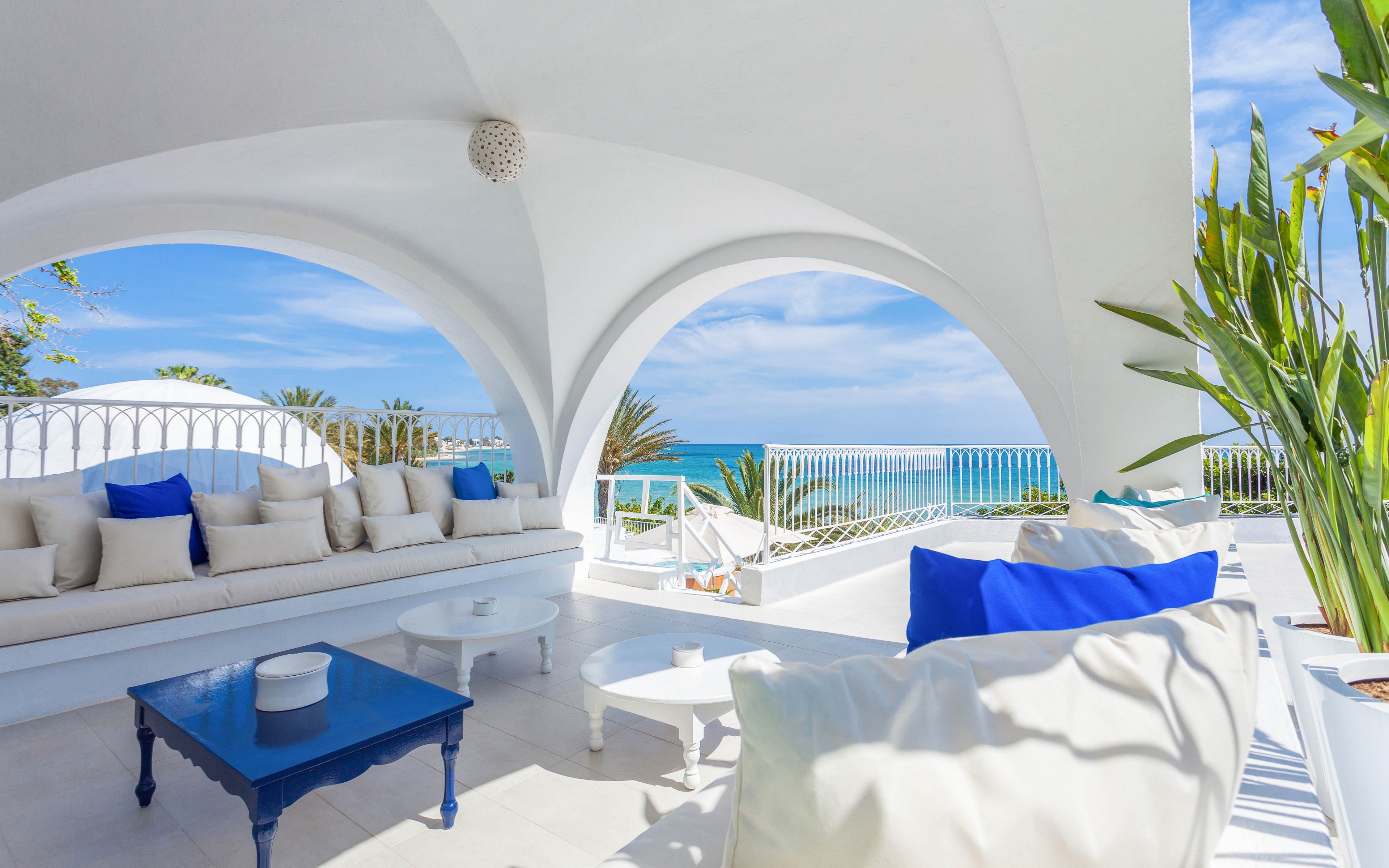 An open-air white terrace lounge features cushioned seating and low tables under elegant arches. Blue accent pillows and a dark blue coffee table contrast with the bright white architecture. The space overlooks a calm blue sea and palm trees beyond a decorative railing. Sunlight and clear skies create a serene, luxurious coastal atmosphere.