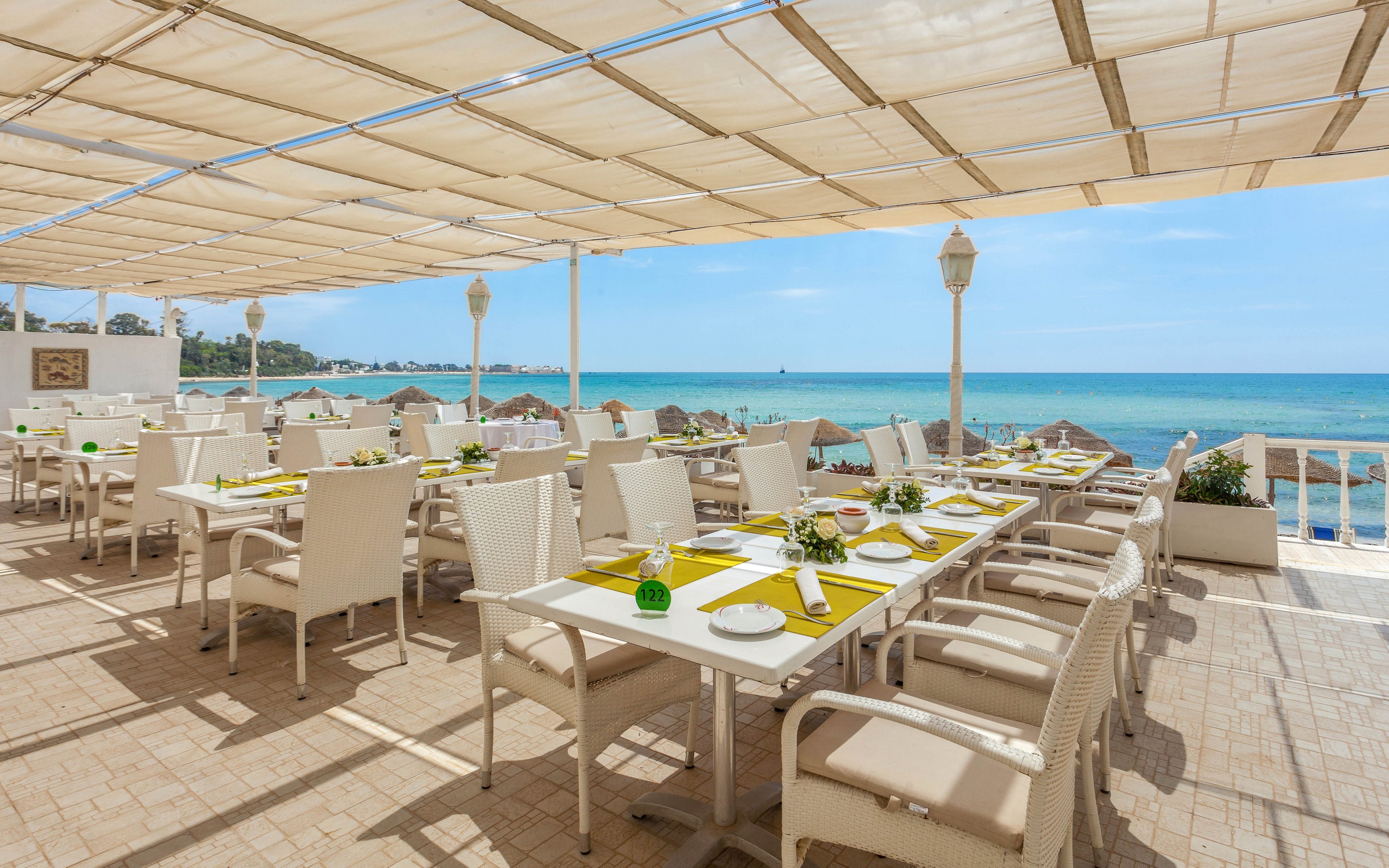 An open-air restaurant terrace overlooks a bright blue ocean on a sunny day. White tables and chairs are neatly arranged under a large canopy that provides shade. Each table is set with plates, glasses, and yellow placemats, with small green decorative accents. The scene conveys a relaxed, coastal dining atmosphere with clear skies and calm water in the background.