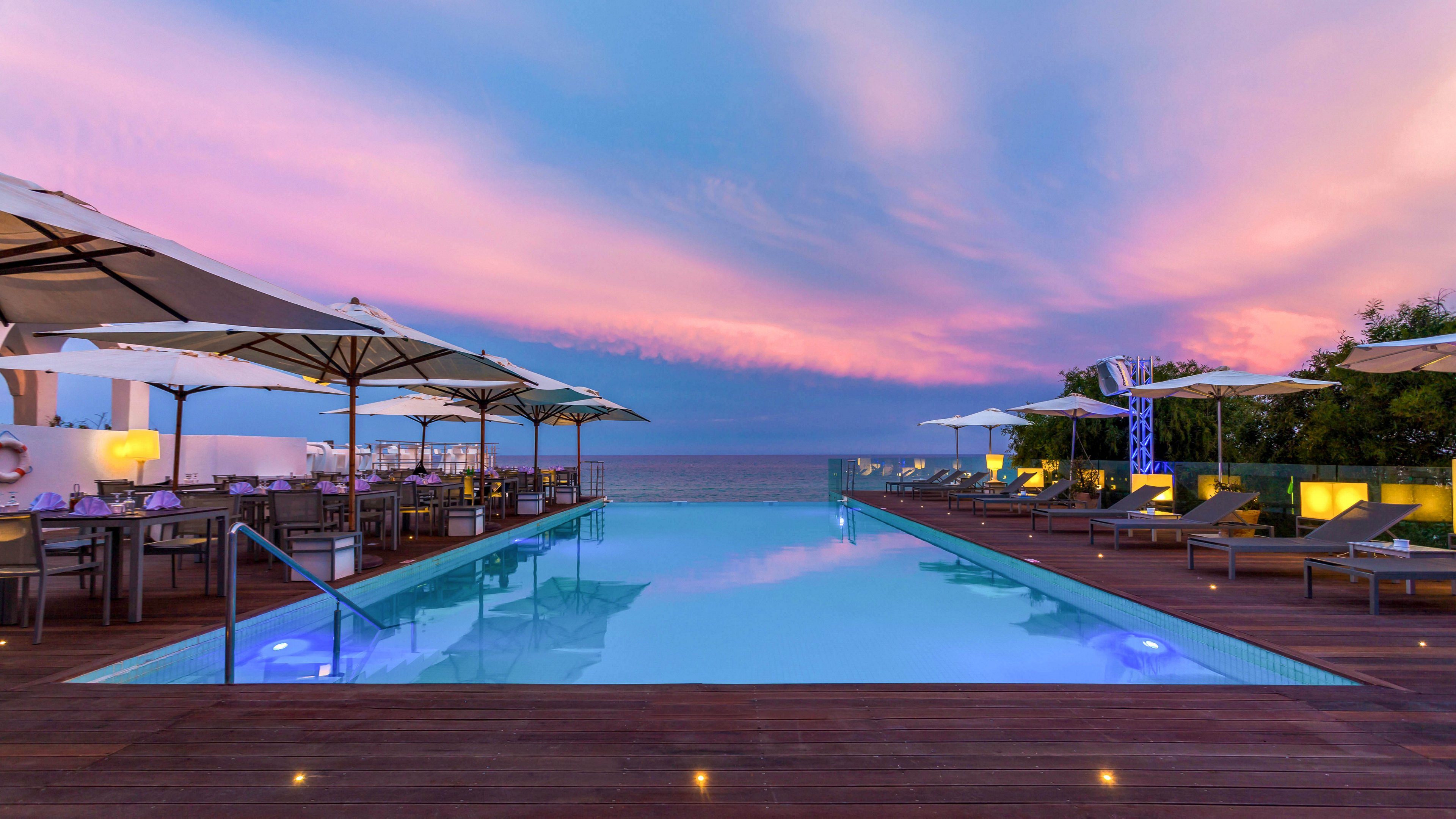 A serene rooftop infinity pool glows with blue lighting under a dramatic sunset sky. Lounge chairs and dining tables with large white umbrellas line both sides of the pool. Soft deck lights and reflections on the water create an elegant, relaxing atmosphere. The horizon and distant sea are visible beyond the glass railing, suggesting a coastal or resort setting.