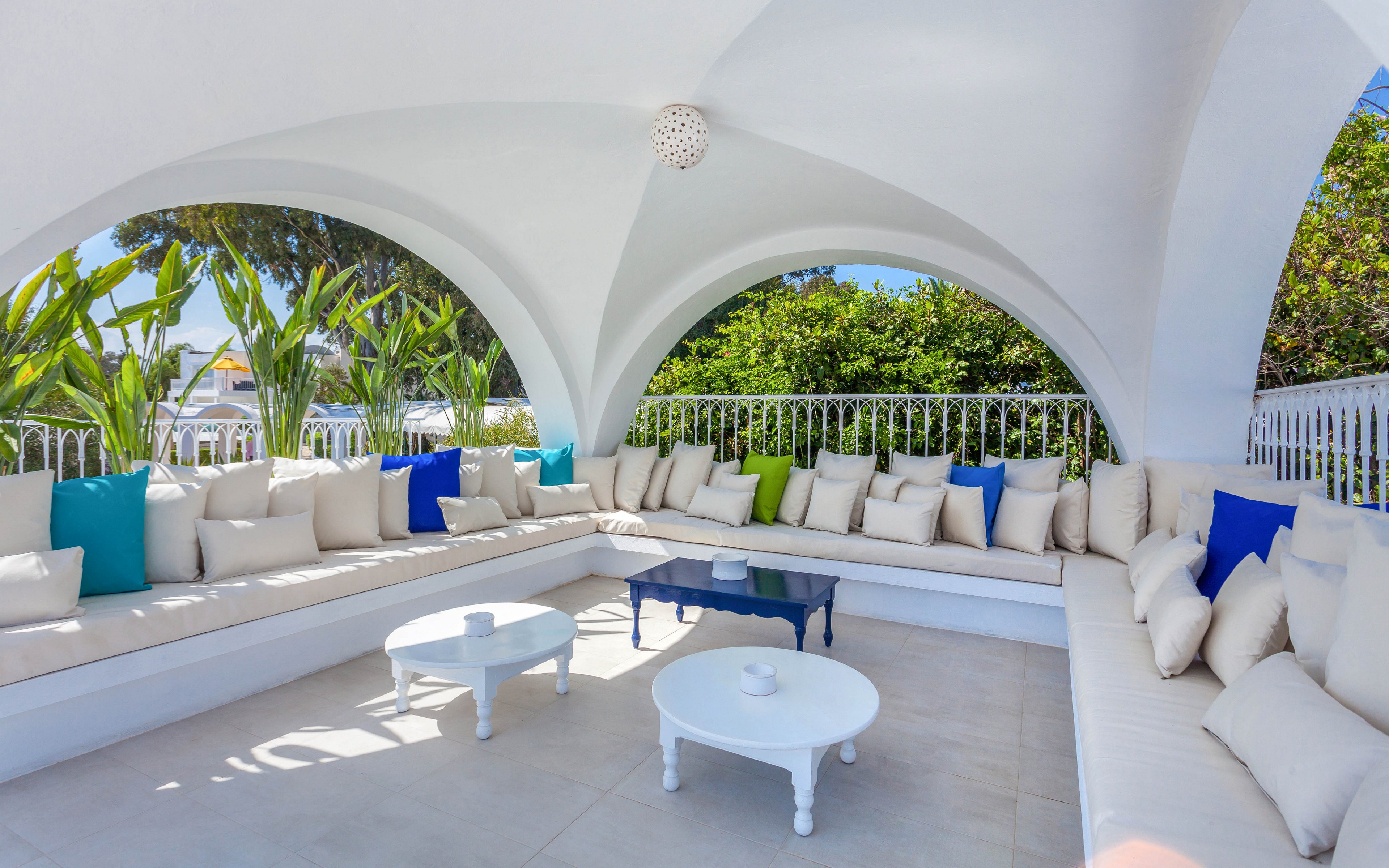 Spacious outdoor lounge area features white built-in seating arranged in a U-shape under smooth white arches. Numerous cream and blue cushions create a relaxed and inviting atmosphere. Two small round tables, one white and one dark blue, sit in the center of the tiled floor. Lush green plants and trees surround the terrace, adding contrast to the bright, sunlit setting.