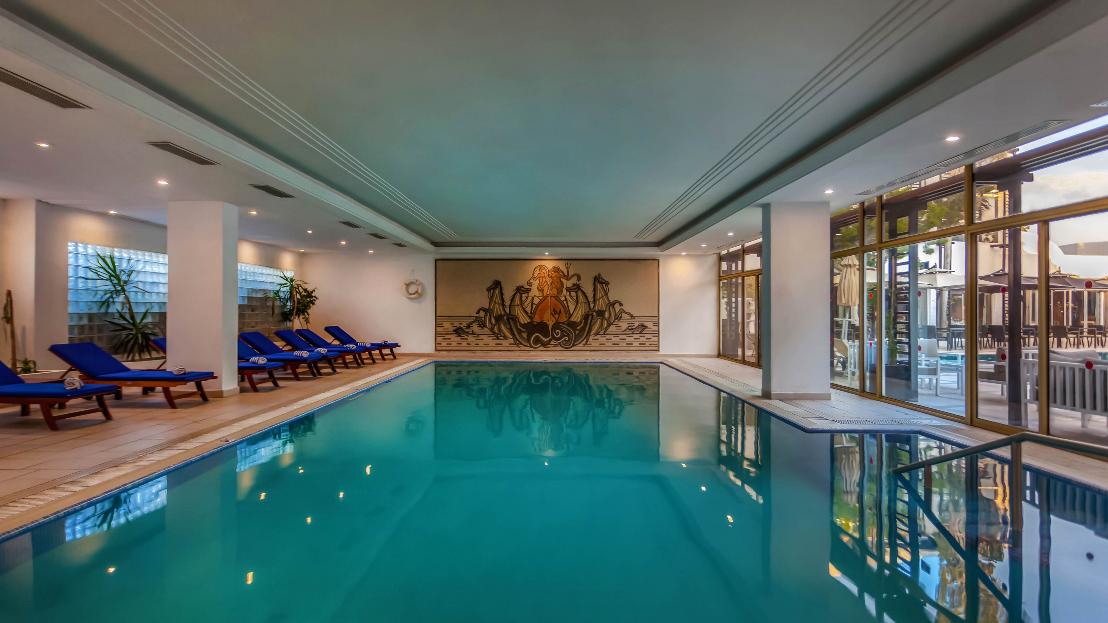Spacious indoor swimming pool inside a modern hotel or spa. Blue water reflects the ceiling lights and large windows along one side of the room. Blue cushioned loungers line the tiled deck, offering a place to relax. A decorative mural with a central figure and ornate design adorns the far wall, adding an artistic focal point.