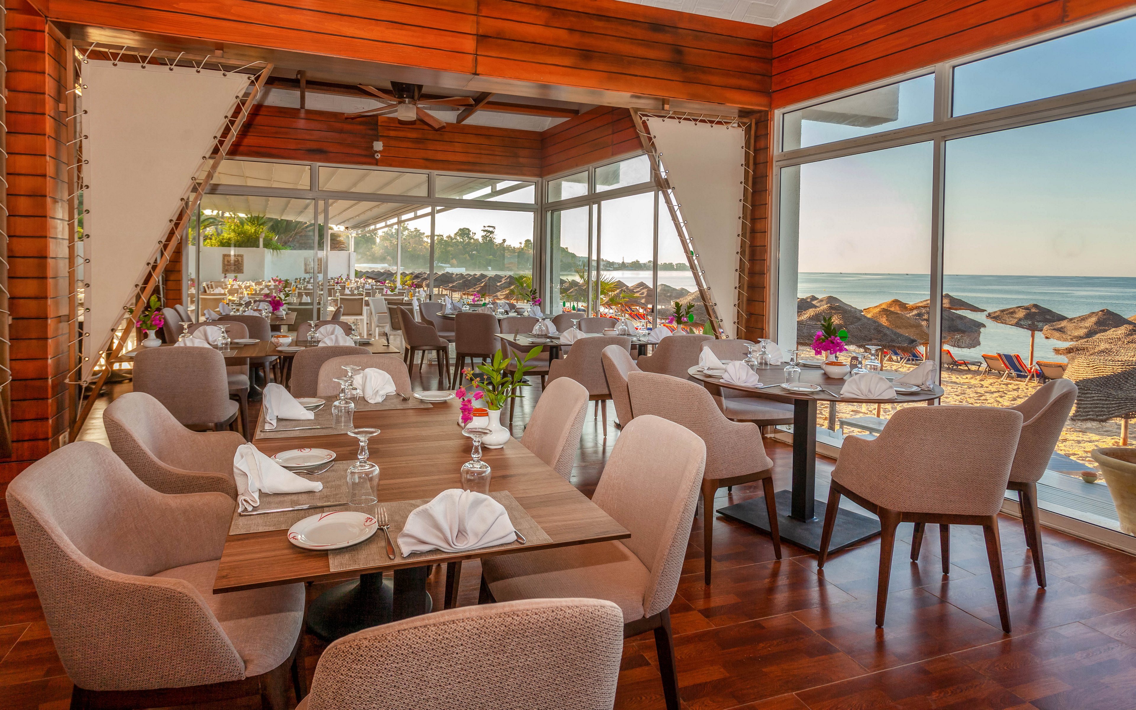 A stylish beachfront restaurant features neatly set wooden tables with folded white napkins and glassware. Soft upholstered chairs surround the tables, creating a comfortable and contemporary dining space. Large floor-to-ceiling windows reveal a sandy beach with sun loungers and umbrellas beside the calm sea. Warm wood paneling and natural light enhance the relaxed, coastal atmosphere.