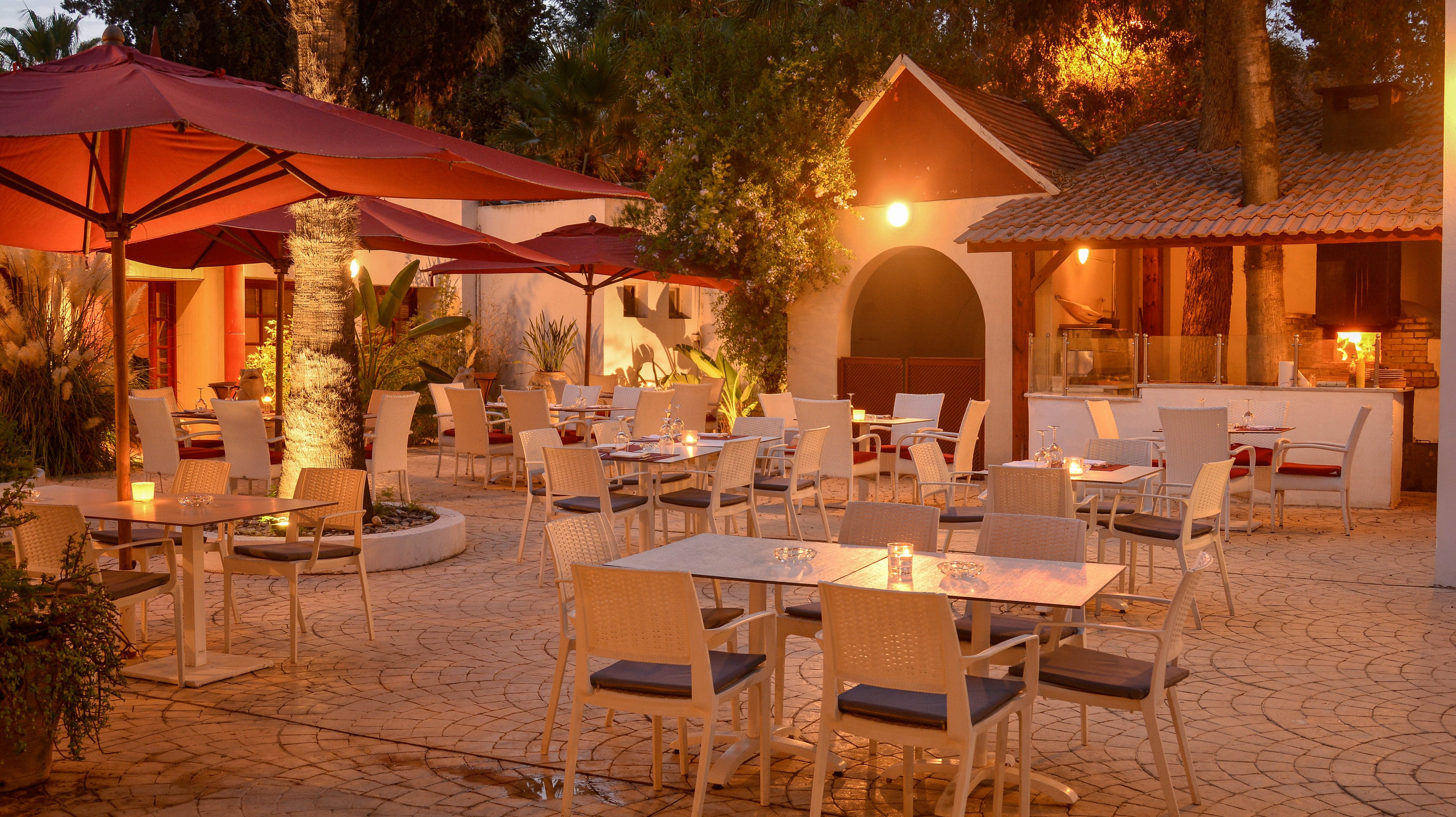 An inviting outdoor restaurant terrace is softly lit at dusk with warm ambient lighting. Empty tables and chairs are neatly arranged on a stone-paved patio, ready for evening diners. Large red umbrellas and surrounding trees create a sheltered, intimate atmosphere. A small building with tiled roof and glowing windows adds to the cozy, Mediterranean-style setting.