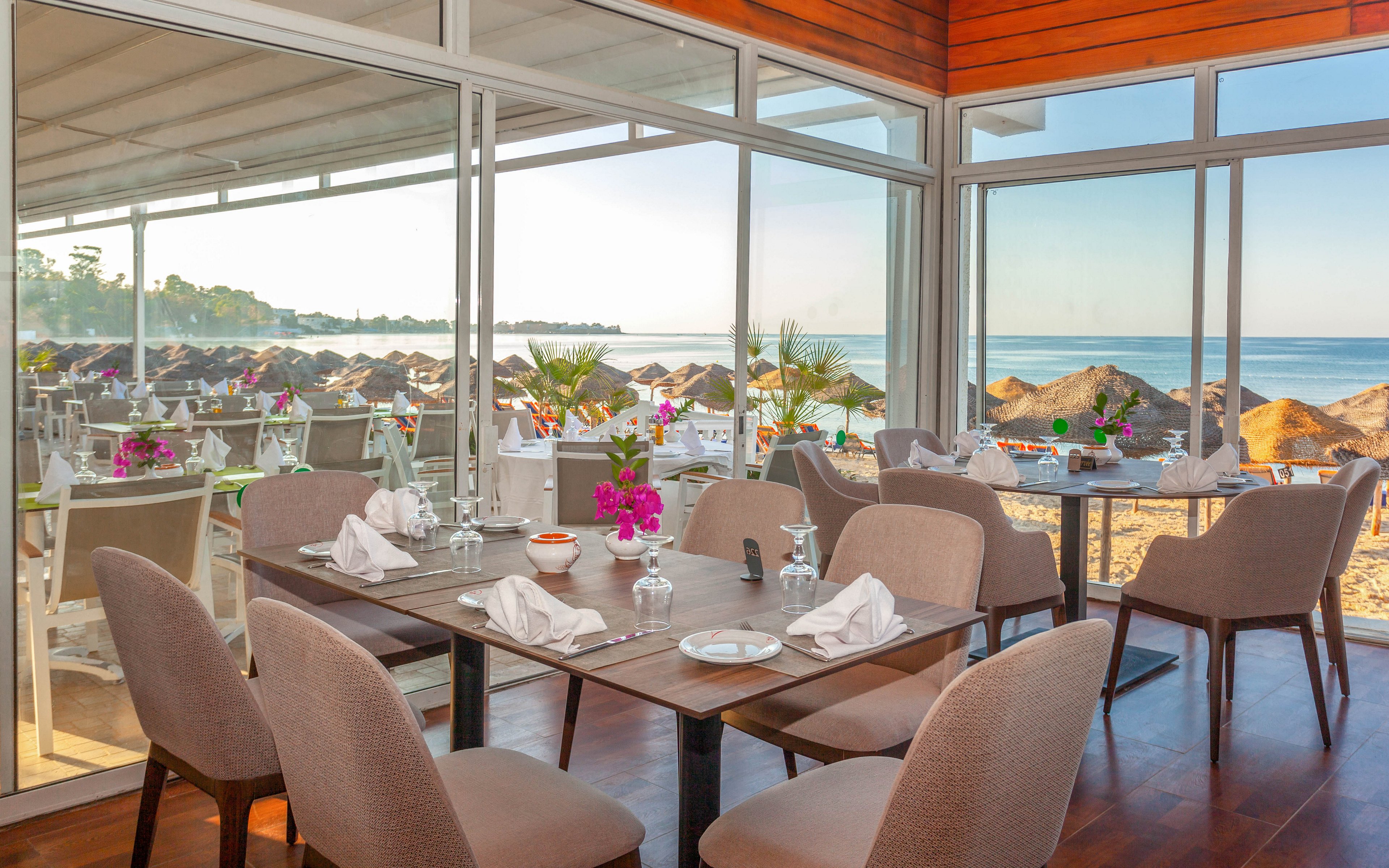 A neatly set restaurant dining area overlooks a sandy beach and calm ocean. Tables are arranged with folded white napkins, glassware, and simple tableware, accented by bright flowers in vases. Large floor-to-ceiling windows and glass doors provide an open view of sun loungers and umbrellas outside. Warm wooden finishes and neutral-toned chairs create a relaxed yet elegant coastal atmosphere.