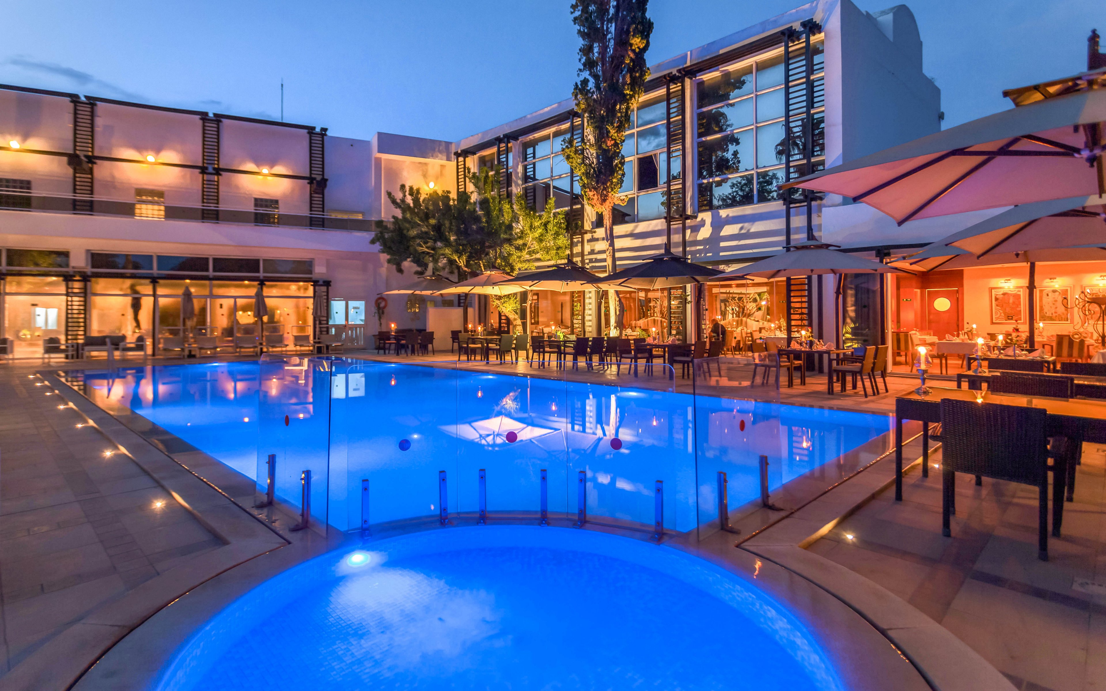 A modern hotel courtyard features a large illuminated swimming pool at dusk. The blue pool lighting contrasts with the warm glow from the surrounding restaurant and lounge areas. Outdoor tables, umbrellas, and seating line the poolside, suggesting a relaxed dining or social atmosphere. The contemporary building facade and clear evening sky enhance the upscale resort ambiance.