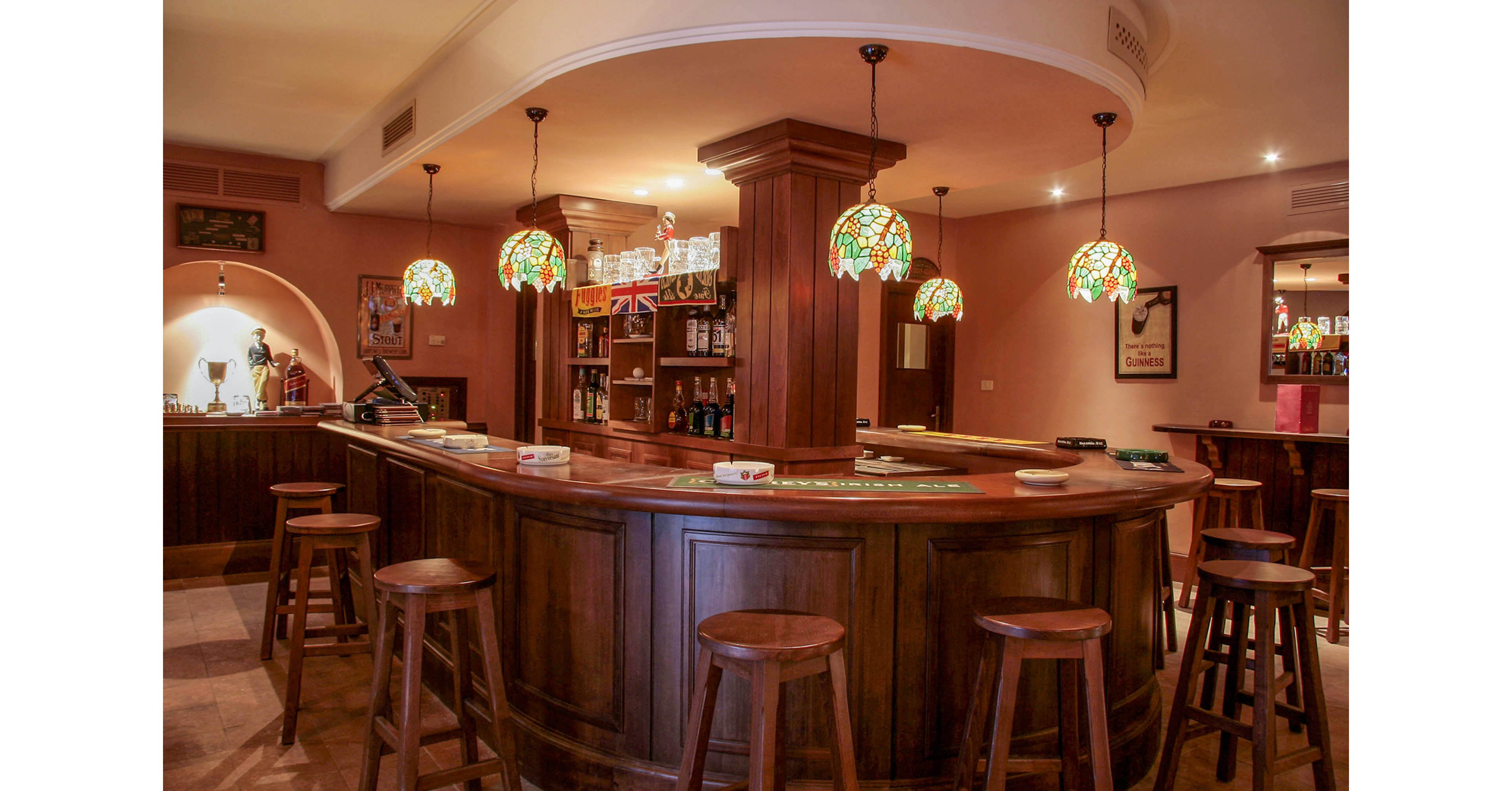 A classic wooden bar counter stands in the center of an empty pub. Stained glass pendant lamps hang above the bar, casting a warm glow over the stools and polished wood surfaces. Shelves in the background display various bottles and glassware, with a small flag adding character to the scene. The overall atmosphere is cozy and inviting, suggesting a quiet moment before guests arrive.