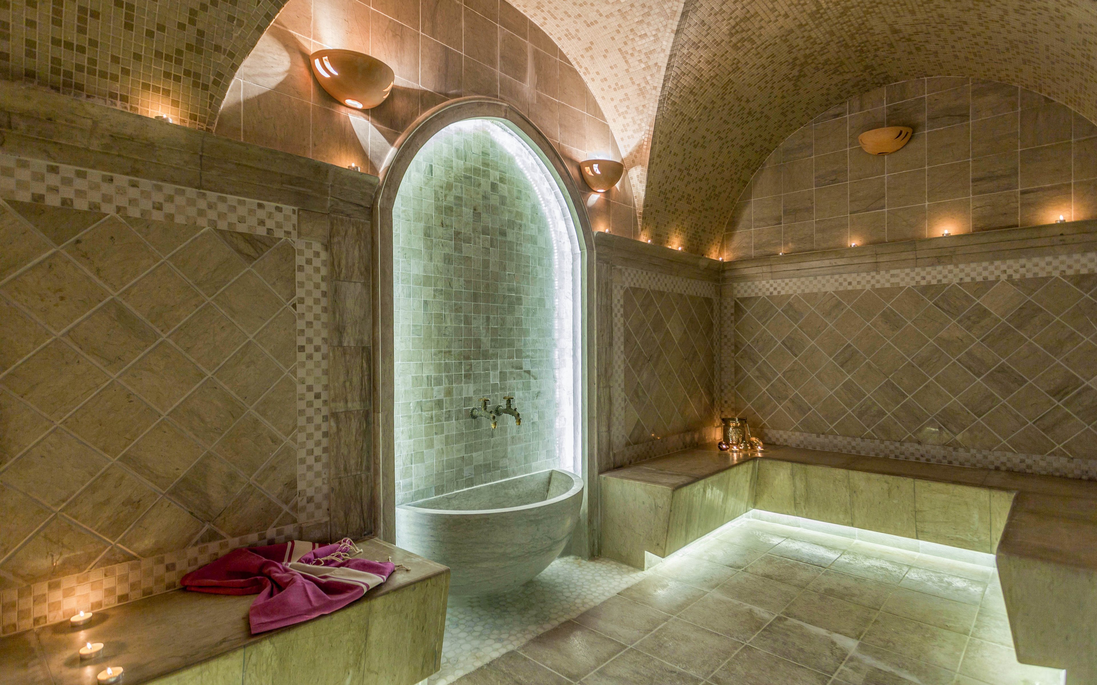 A traditional stone hammam interior is softly illuminated by candles and hidden floor lighting. An arched shower alcove with a stone basin forms the focal point of the room. Long stone benches line the walls, creating a serene space for relaxation. A folded pink towel and small items rest on one of the benches, adding a touch of color to the warm, neutral tiles.