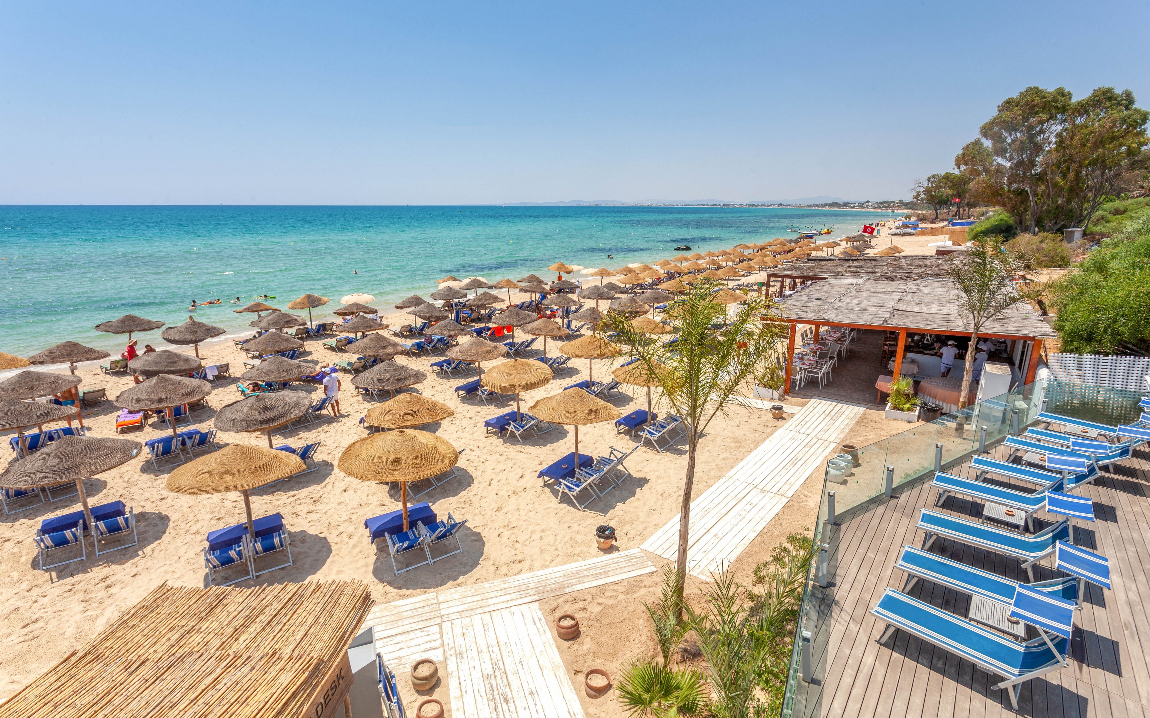A crowded sandy beach is lined with rows of straw umbrellas and blue sun loungers facing the turquoise sea. A wooden boardwalk leads from the resort area down to the shoreline, where people relax and sunbathe. On the right, a raised deck with additional loungers overlooks the beach and a small open-air bar or restaurant. The clear sky and calm water create a bright, summery vacation atmosphere.