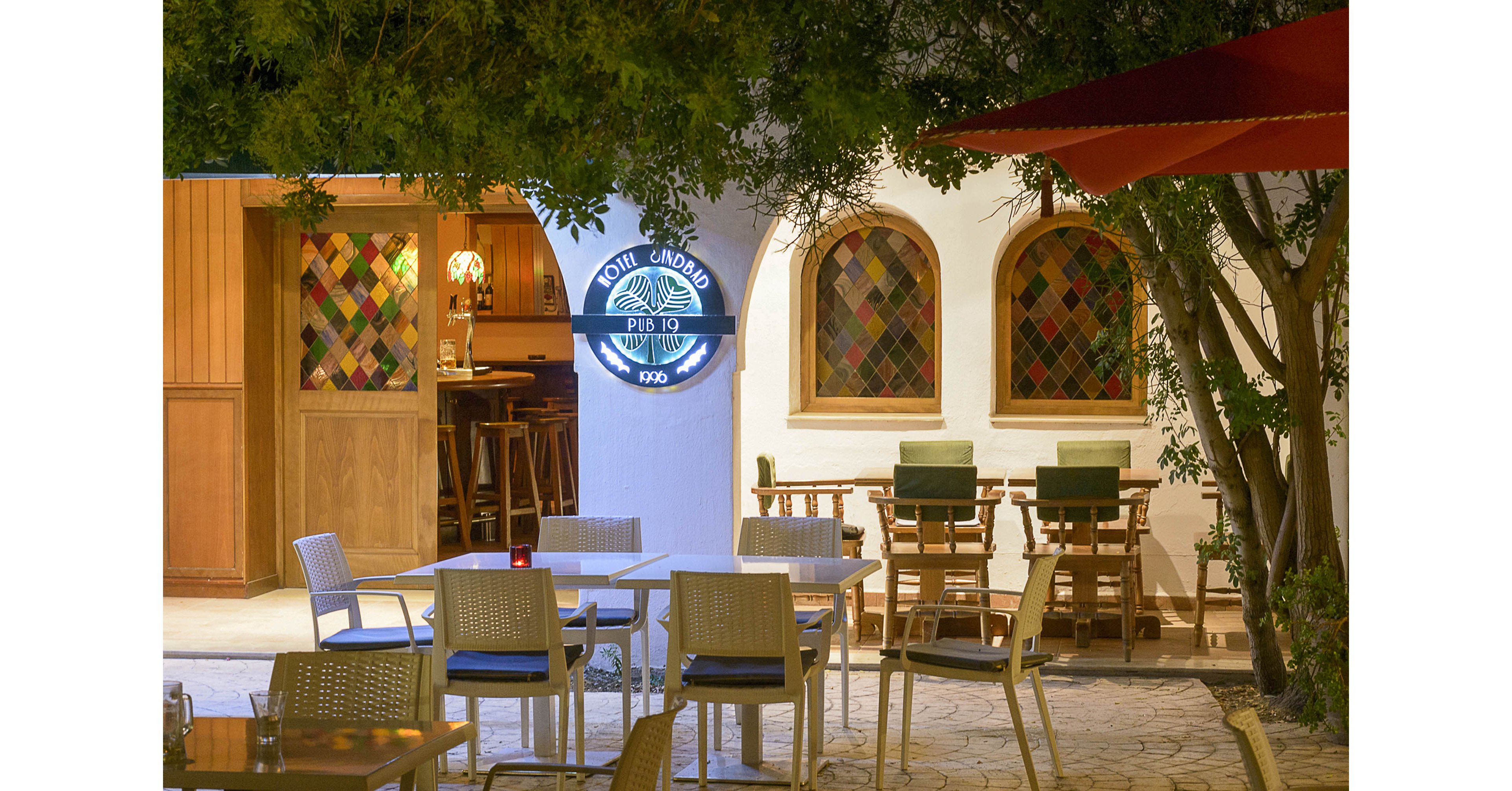 An inviting restaurant terrace is shown at night with empty tables and chairs arranged on a stone patio. Warm light spills from the interior bar area, where colorful stained-glass windows add a decorative touch. A round illuminated sign with text is mounted on the white wall near the entrance. Trees and large umbrellas frame the seating area, creating a relaxed, intimate atmosphere.